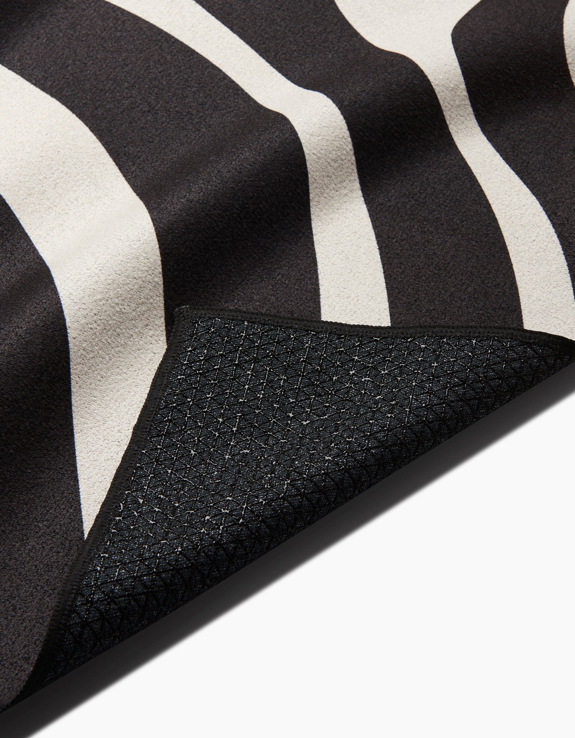 Close-up of GEOMETRYs Spiral slip-resistant yoga towel, featuring a black and white wavy stripe pattern. A flipped corner reveals its textured black underside with a subtle grid design, set against a white background.
