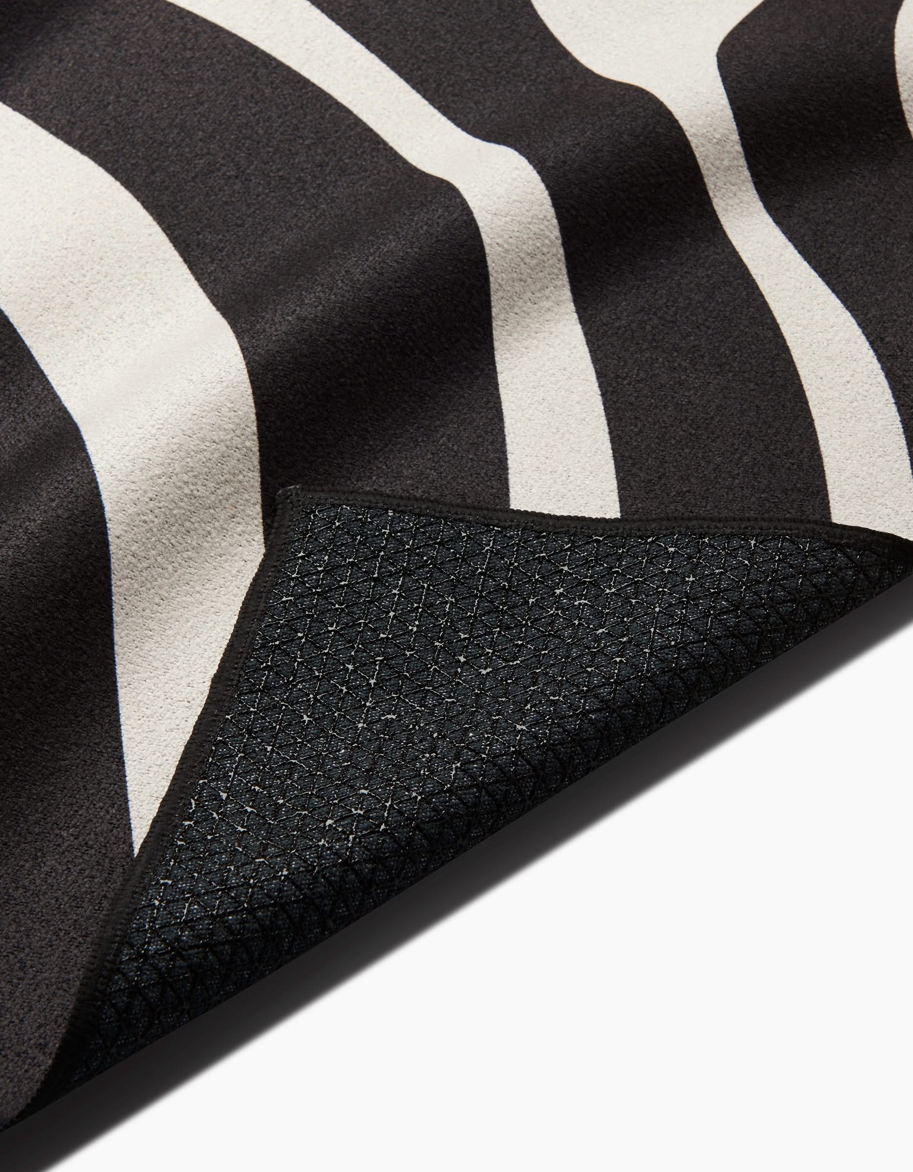 Close-up of GEOMETRYs Spiral slip-resistant yoga towel, featuring a black and white wavy stripe pattern. A flipped corner reveals its textured black underside with a subtle grid design, set against a white background.