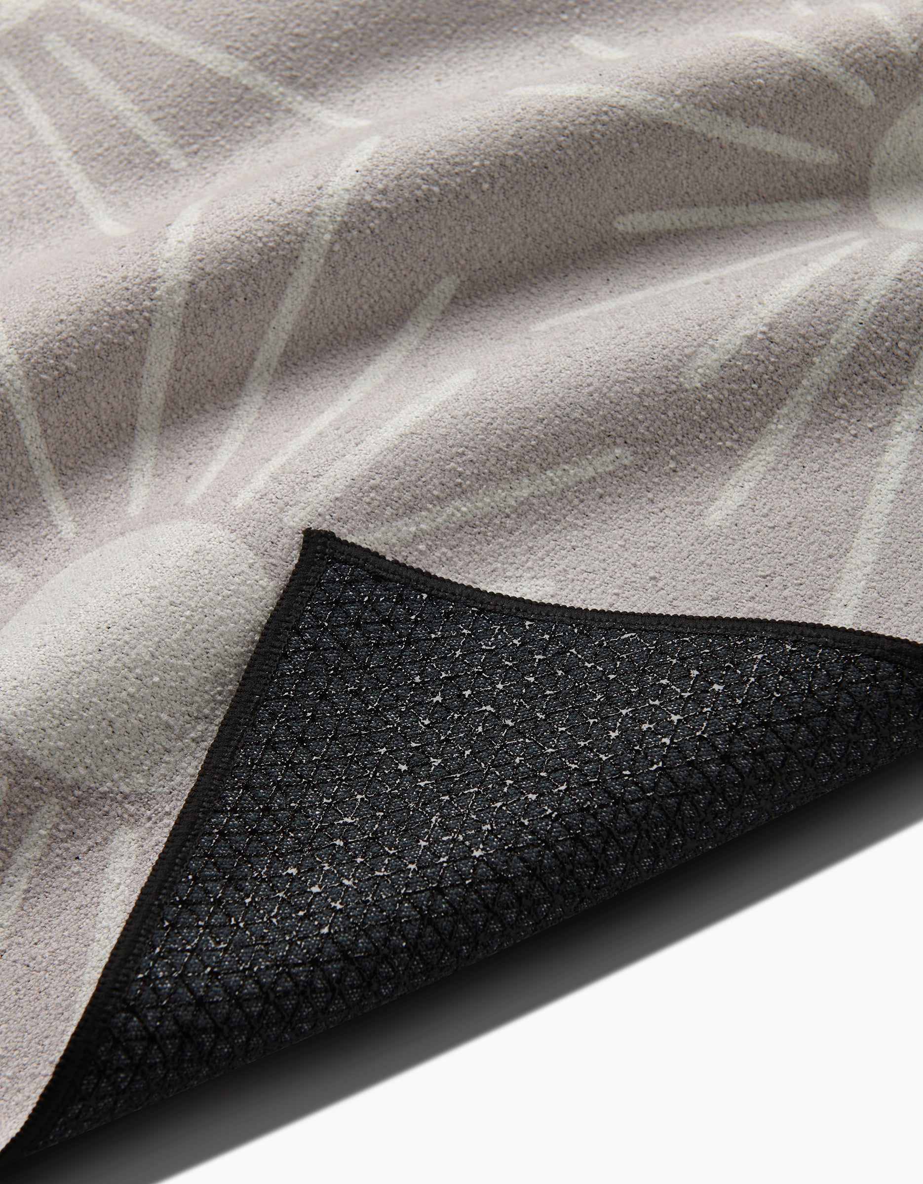 A close-up of the Hufton Studio Sunny Geo yoga towel shows its textured beige surface with a white abstract sunburst design. The flipped corner reveals a black, non-slip silicone grip backing featuring a geometric pattern.