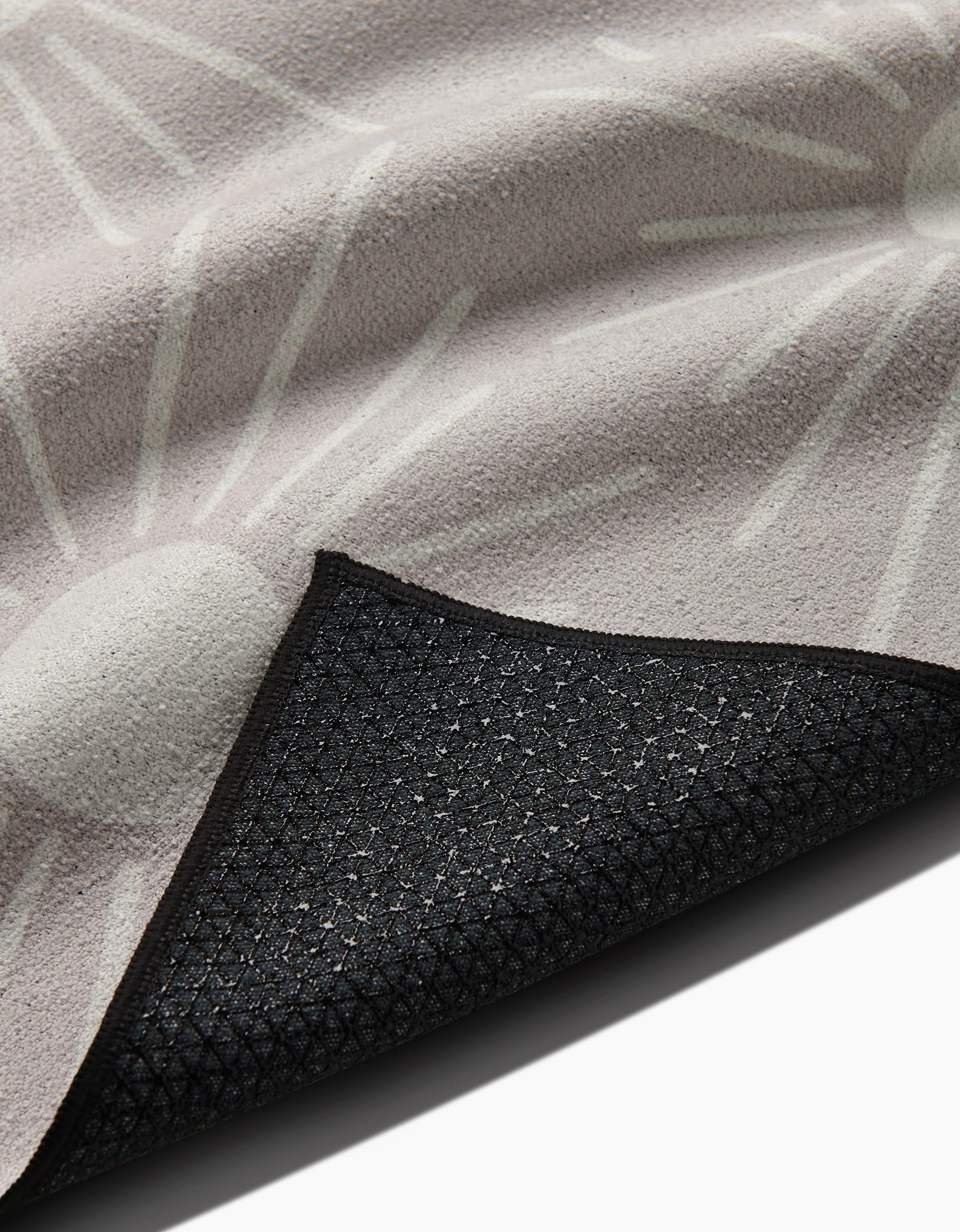 A close-up of the Hufton Studio Sunny Geo yoga towel shows its textured beige surface with a white abstract sunburst design. The flipped corner reveals a black, non-slip silicone grip backing featuring a geometric pattern.