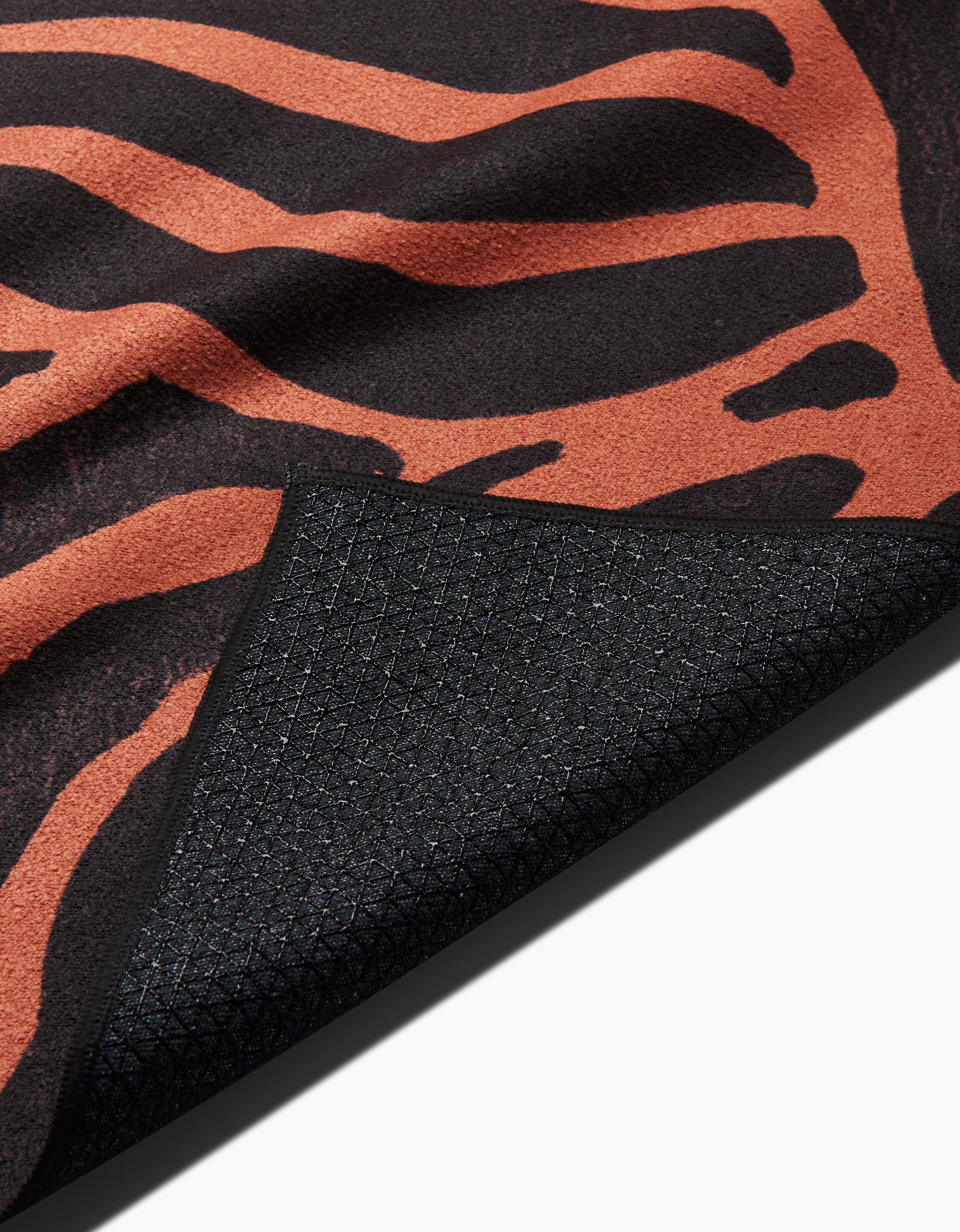 The GEOMETRY Tiger yoga towel features an orange and black tiger stripe pattern, with an upturned corner revealing its slip-resistant, textured underside made from recycled materials.