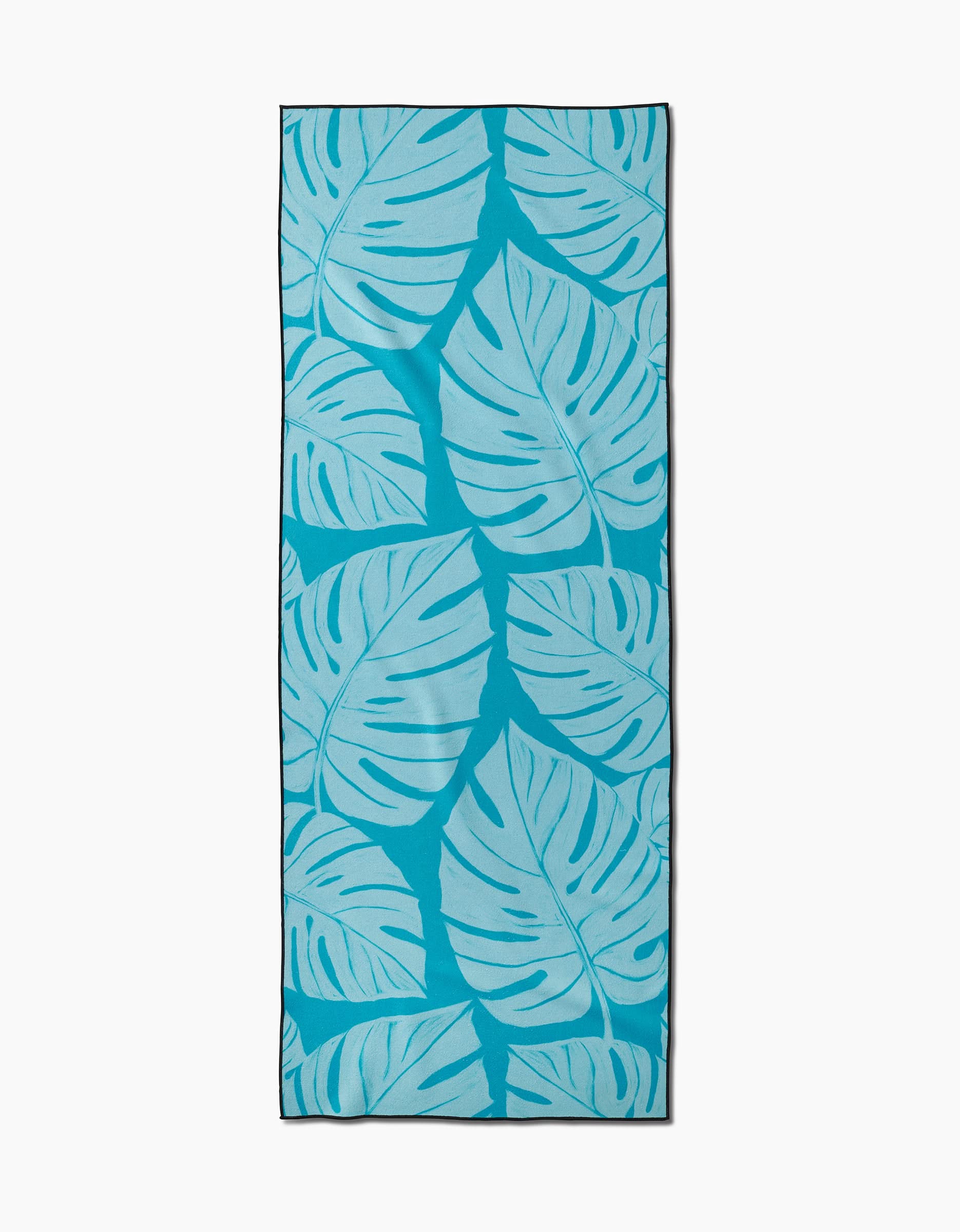 The GEOMETRY Tropical yoga towel is rectangular, blue, and non-slip, featuring a repeating pattern of large overlapping light blue tropical leaves. Shown flat on a white background.
