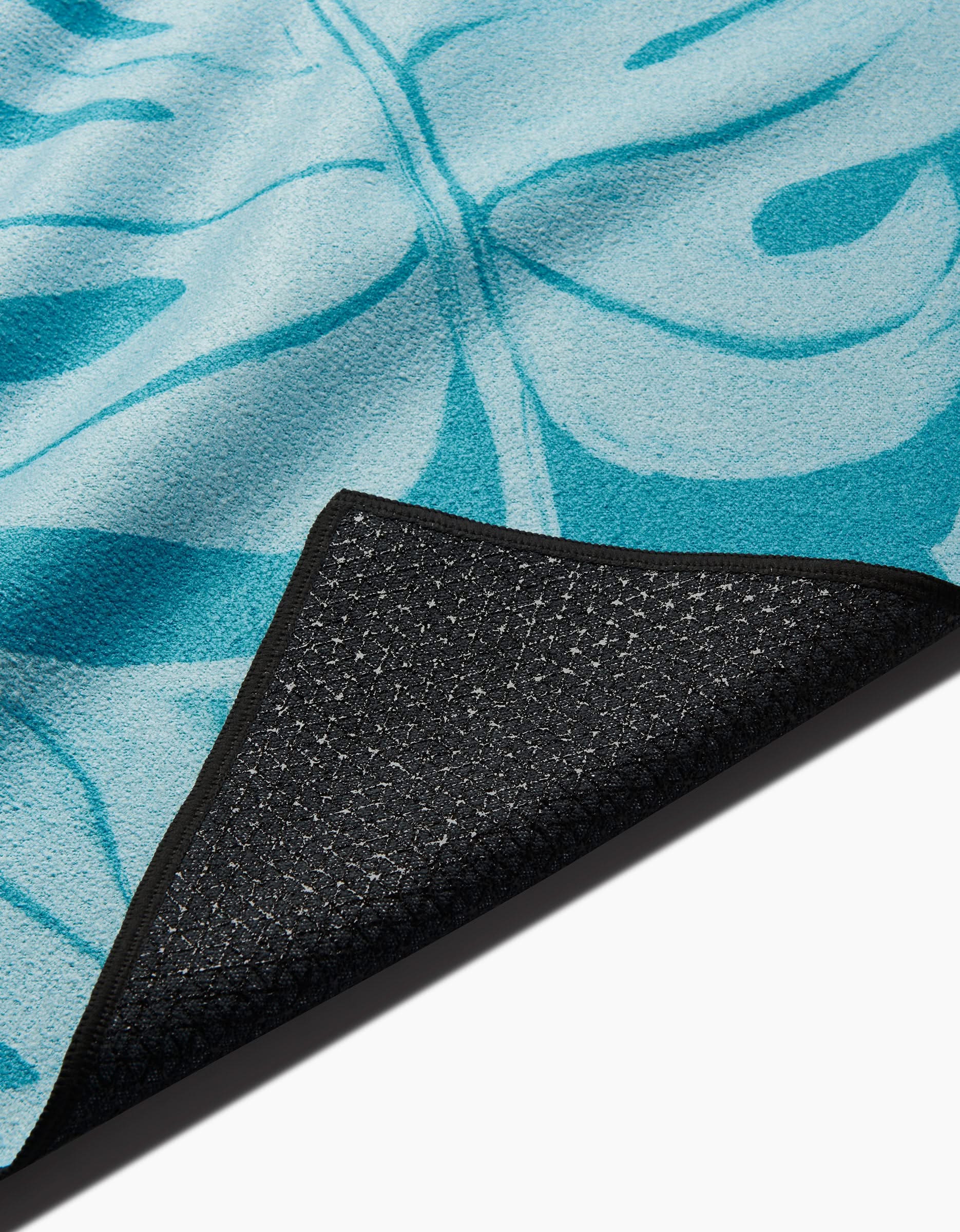 The GEOMETRY Tropical yoga towel, featuring a blue leaf pattern and non-slip textured black backing, is displayed flat with one corner flipped up.