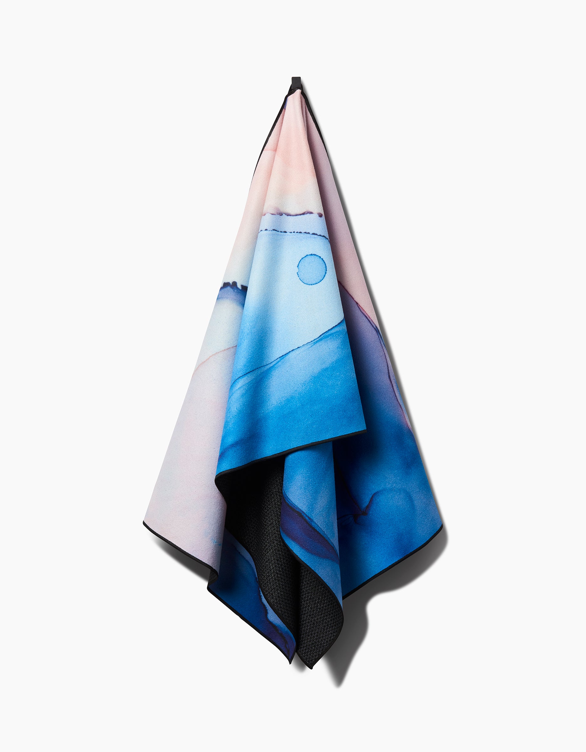 The GEOMETRY Water And Ink yoga towel, designed with a blue and pink abstract mountain landscape, hangs from a hook against a white background. It features slip-resistant material for added stability.