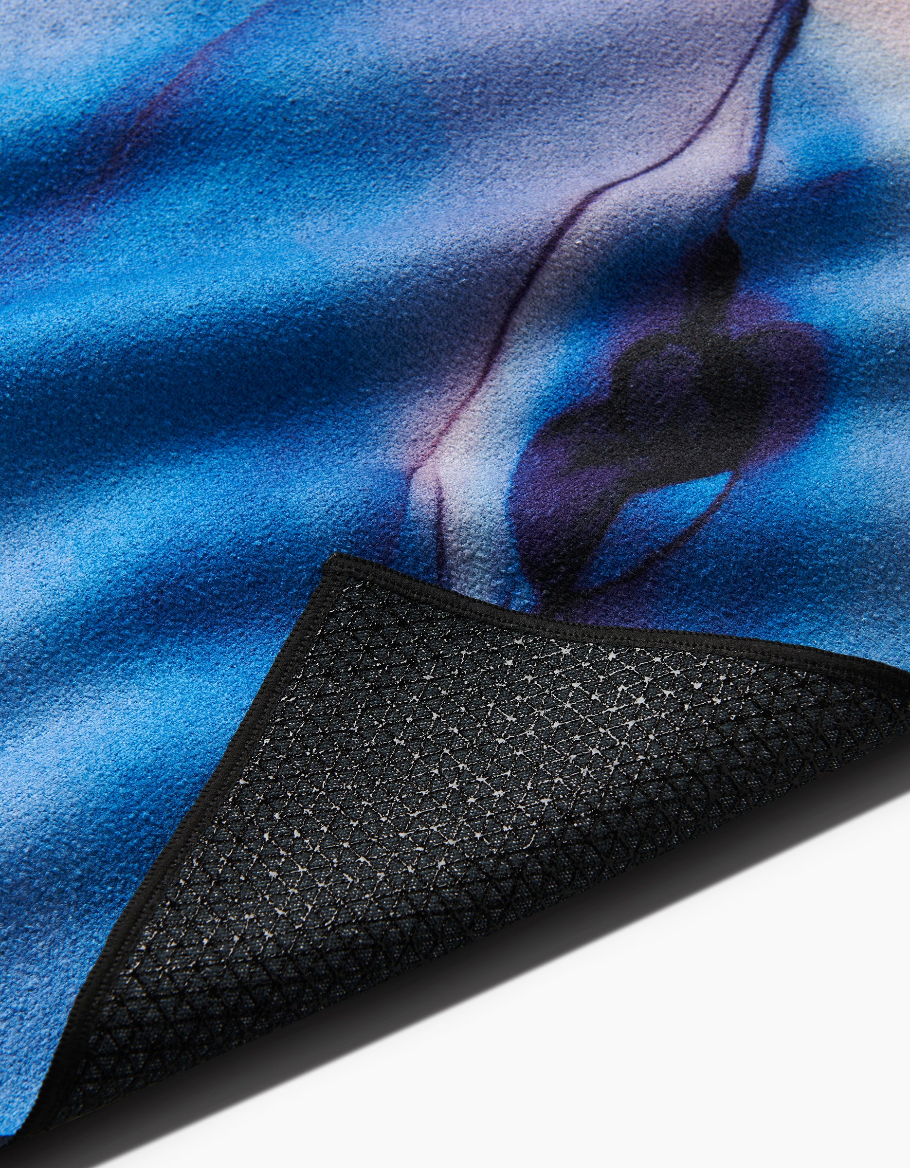 A close-up of GEOMETRYs Water And Ink yoga towel, featuring a blue and purple pattern, soft texture, and a partially folded view showing its black, recycled slip-resistant underside.