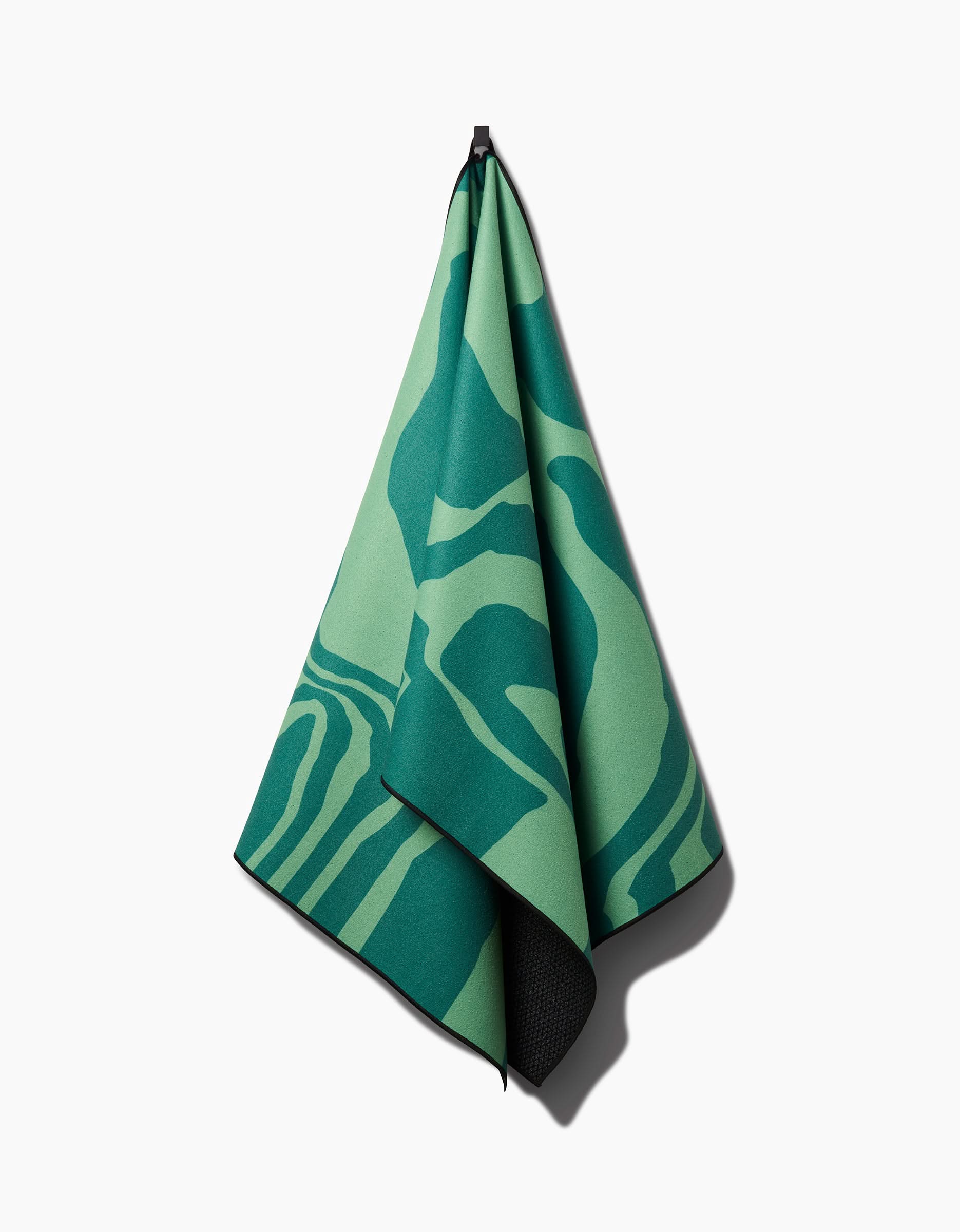 The GEOMETRY Watermelon Vibes yoga towel, featuring abstract wavy green patterns and a silicone grip, hangs from a black hook against a plain white background.