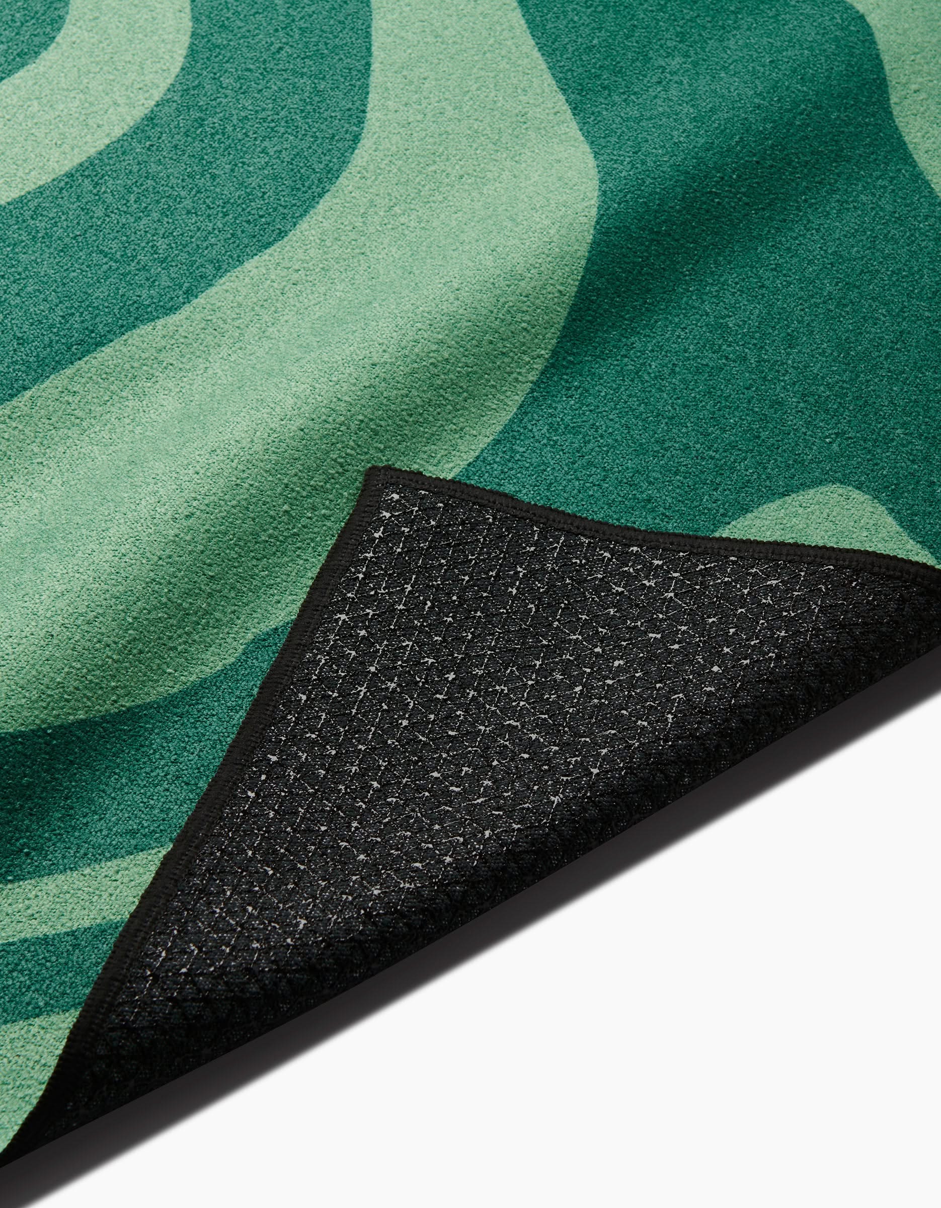 A close-up of the GEOMETRY Watermelon Vibes rug, featuring green wavy patterns and a flipped corner that reveals its black, textured non-slip backing made from recycled materials.