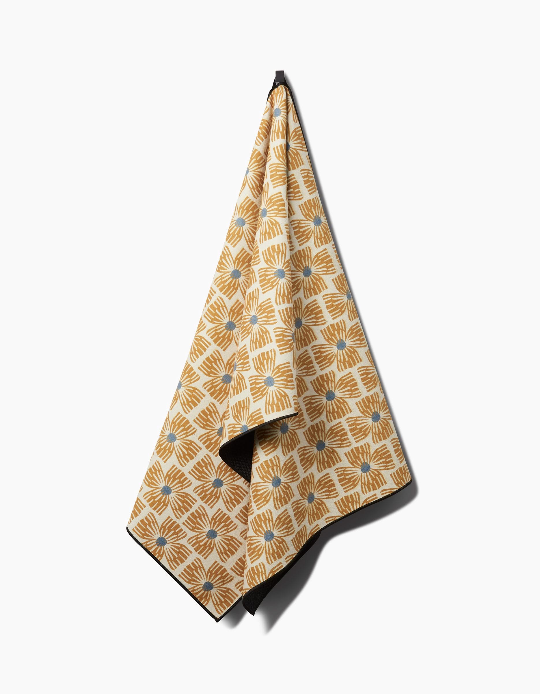 The Ash Cascade Wildflower yoga towel, featuring a yellow and white geometric floral pattern with blue accents and made from recycled materials, hangs on a hook against a plain white background.