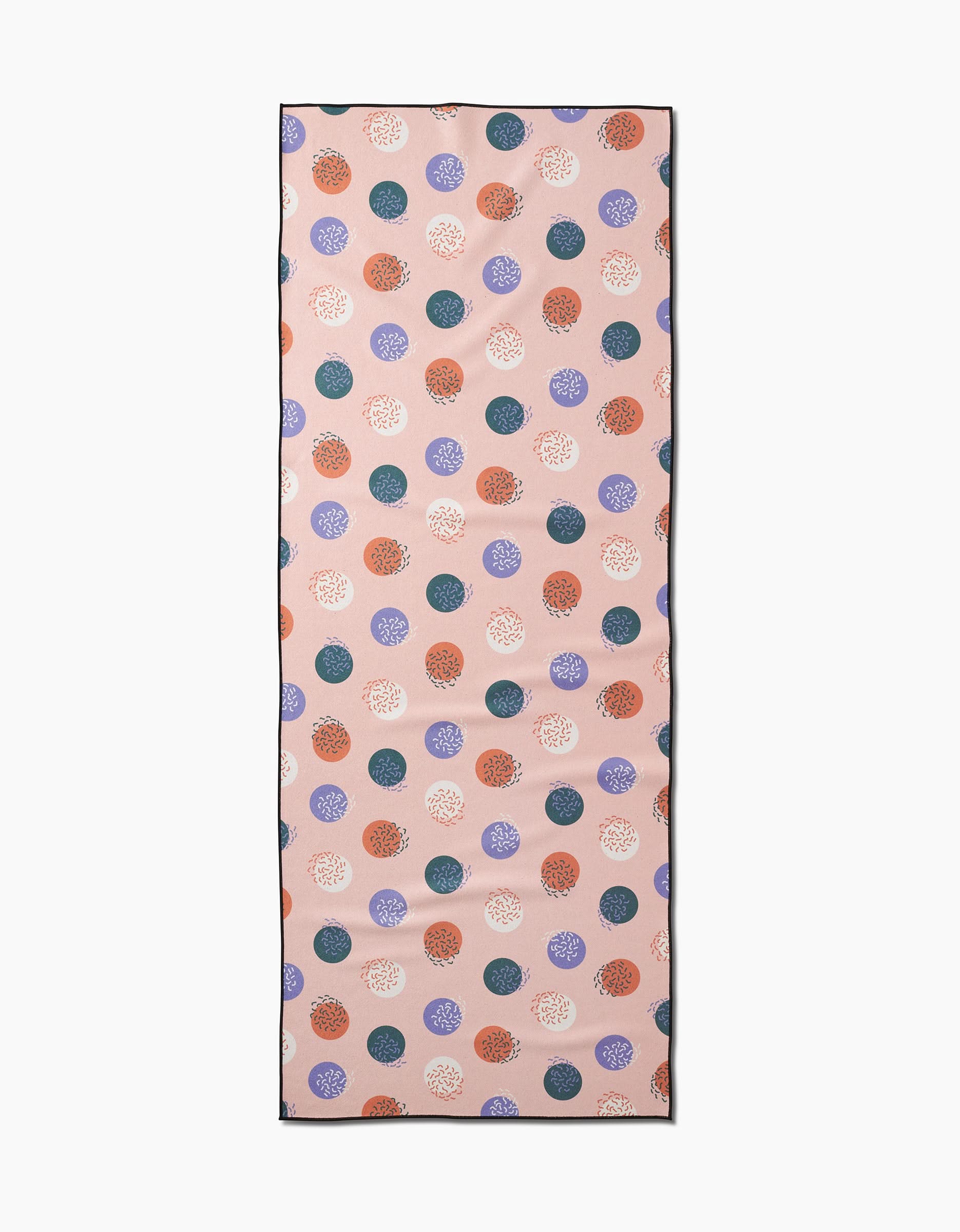 The Wonky Dots yoga towel by Claire Iglesias features a rectangular recycled design with a light pink background and scattered blue, orange, purple, and white polka dots, each adorned with intricate circular patterns.