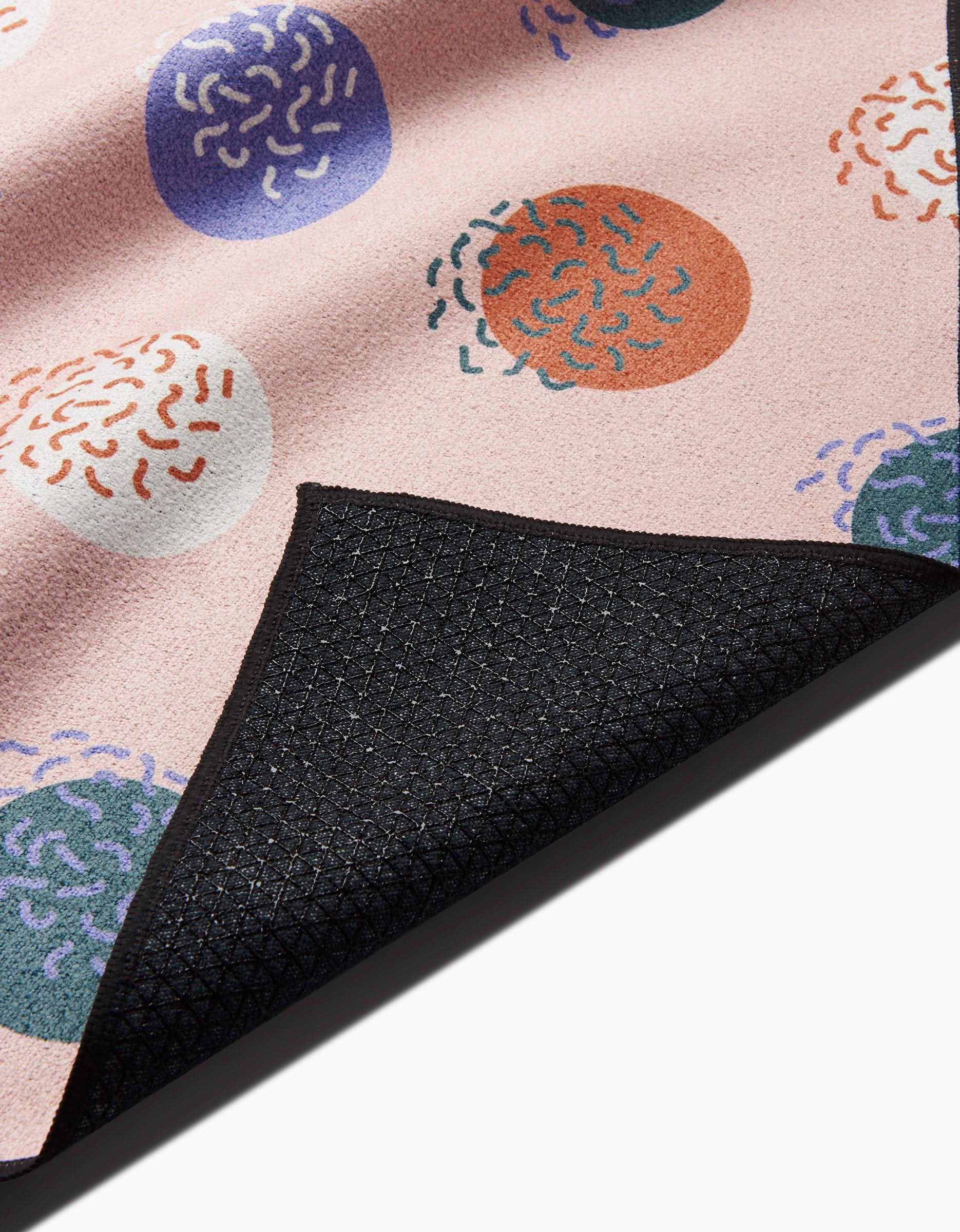 A close-up of the Claire Iglesias Wonky Dots yoga towel shows its soft pink recycled fabric with colorful dots and abstract squiggles, plus a black textured corner flipped over to reveal the underside.