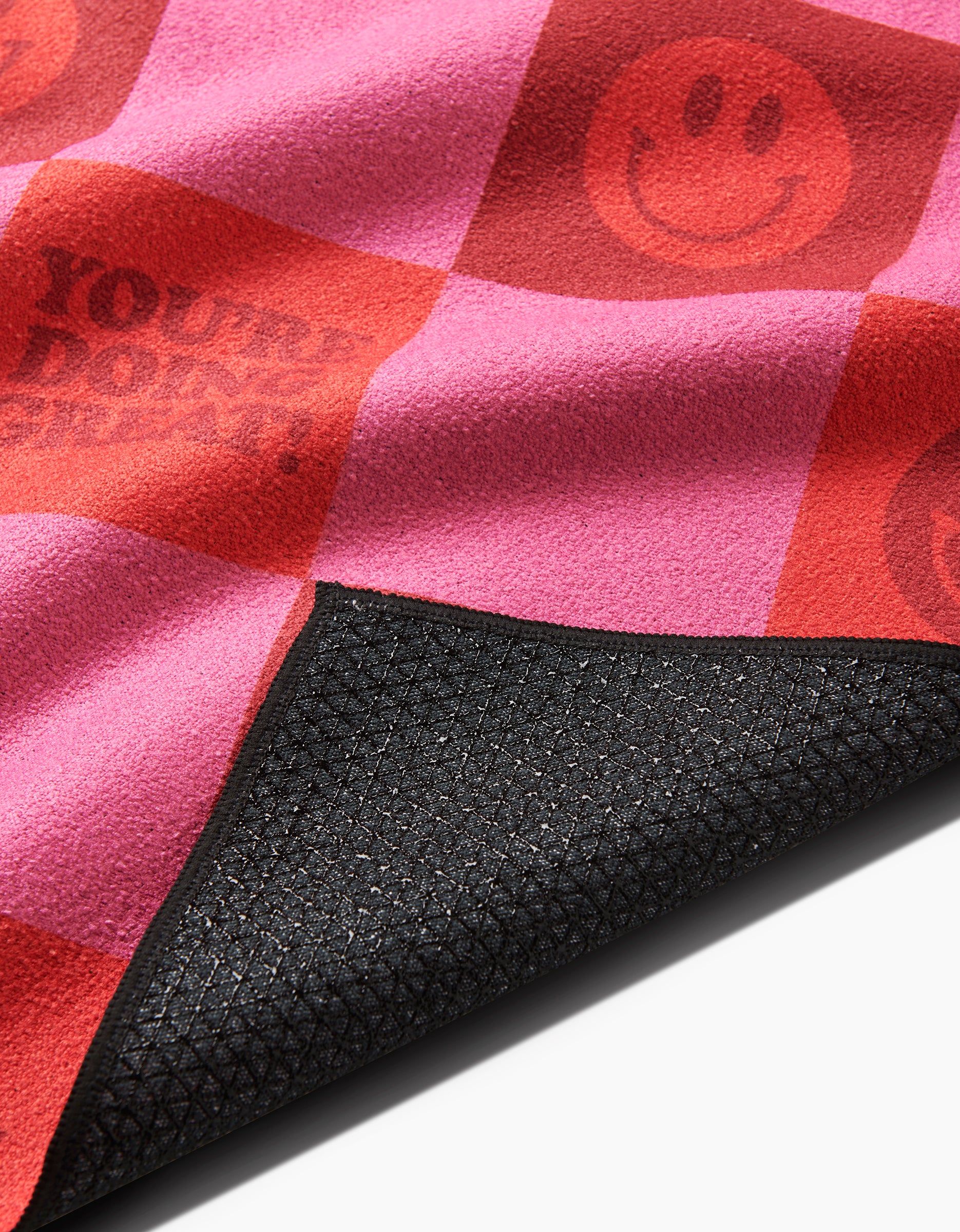 A close-up of the Youre Doing Great yoga towel by Quotes by Christie, featuring pink and red checkers with smiley faces, partial text, and a black textured silicone grip underside at one corner.