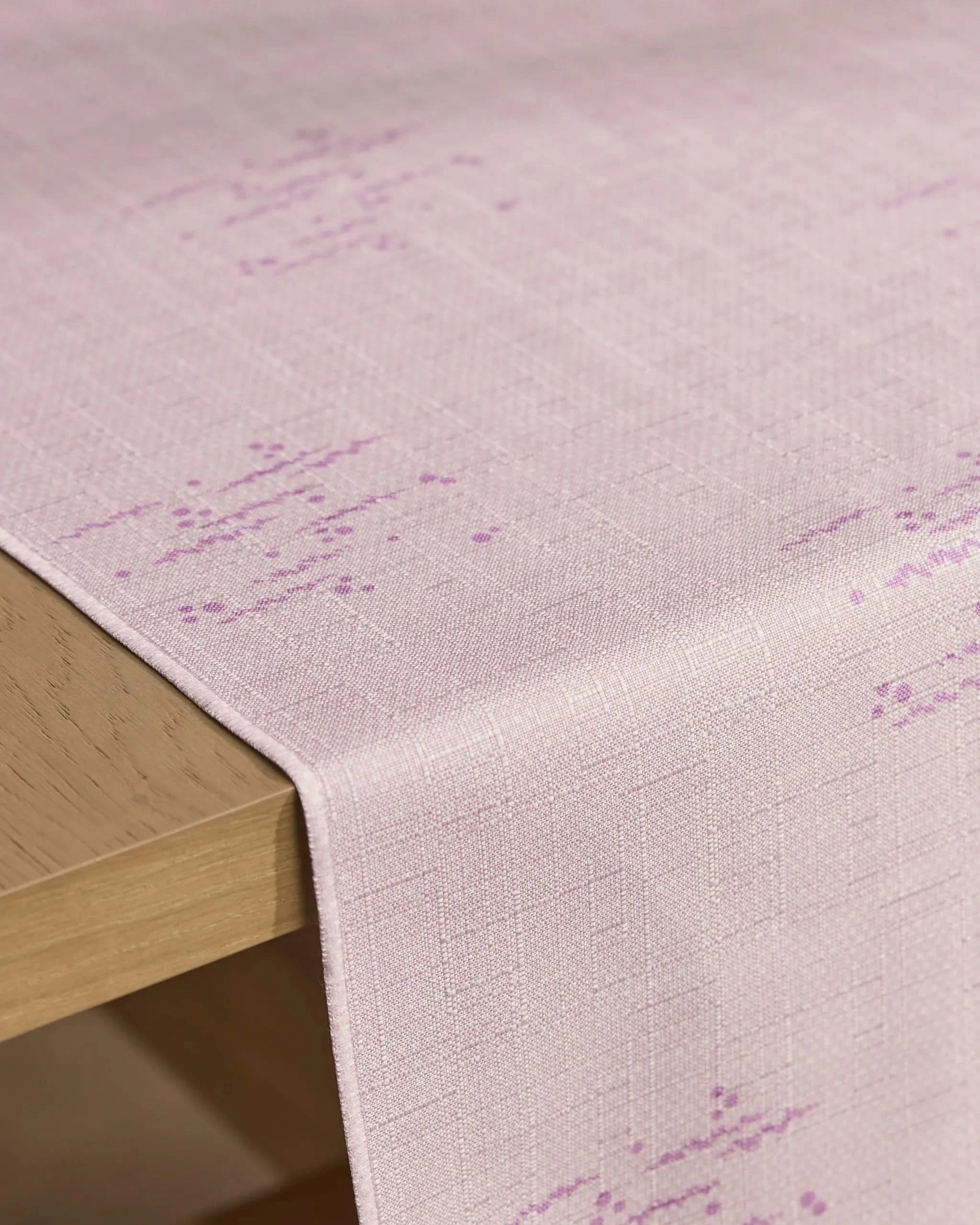 The Geometry Bubble Ditzy Dusty Rose table runner, featuring a light pink stain-resistant fabric with a subtle textured pattern and faint purple accents, drapes over the edge of a light wooden table.