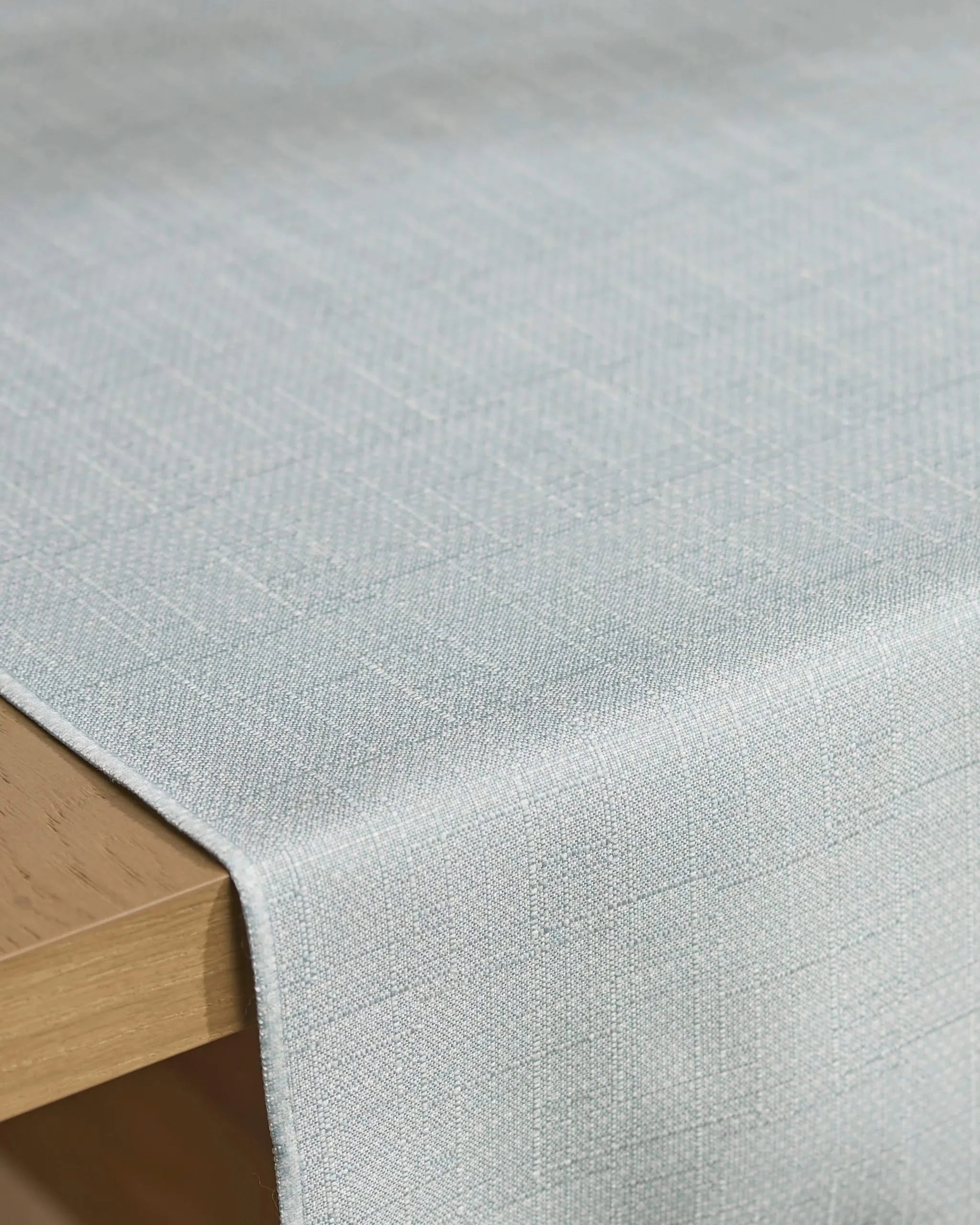 The Geometry Chambray Light Blue table runner, made from recycled materials, drapes over a wooden table and features a subtle checkered woven pattern.