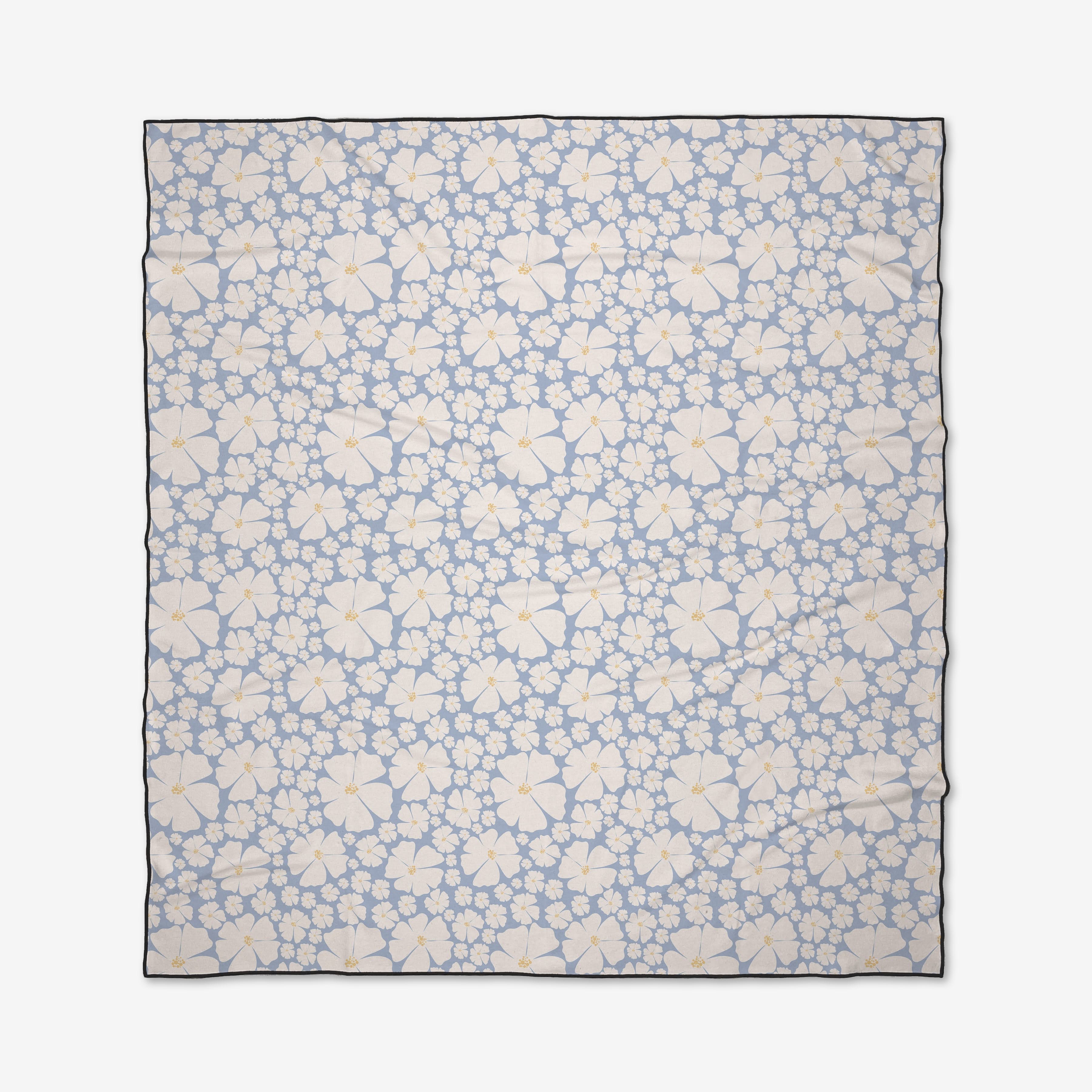 The Daisy Girl Blue beach blanket from Charlie Rowan Designs features a light blue background with white and light yellow hibiscus flowers with orange centers. Quick-drying and made from recycled materials, it’s finished with a thin dark border.