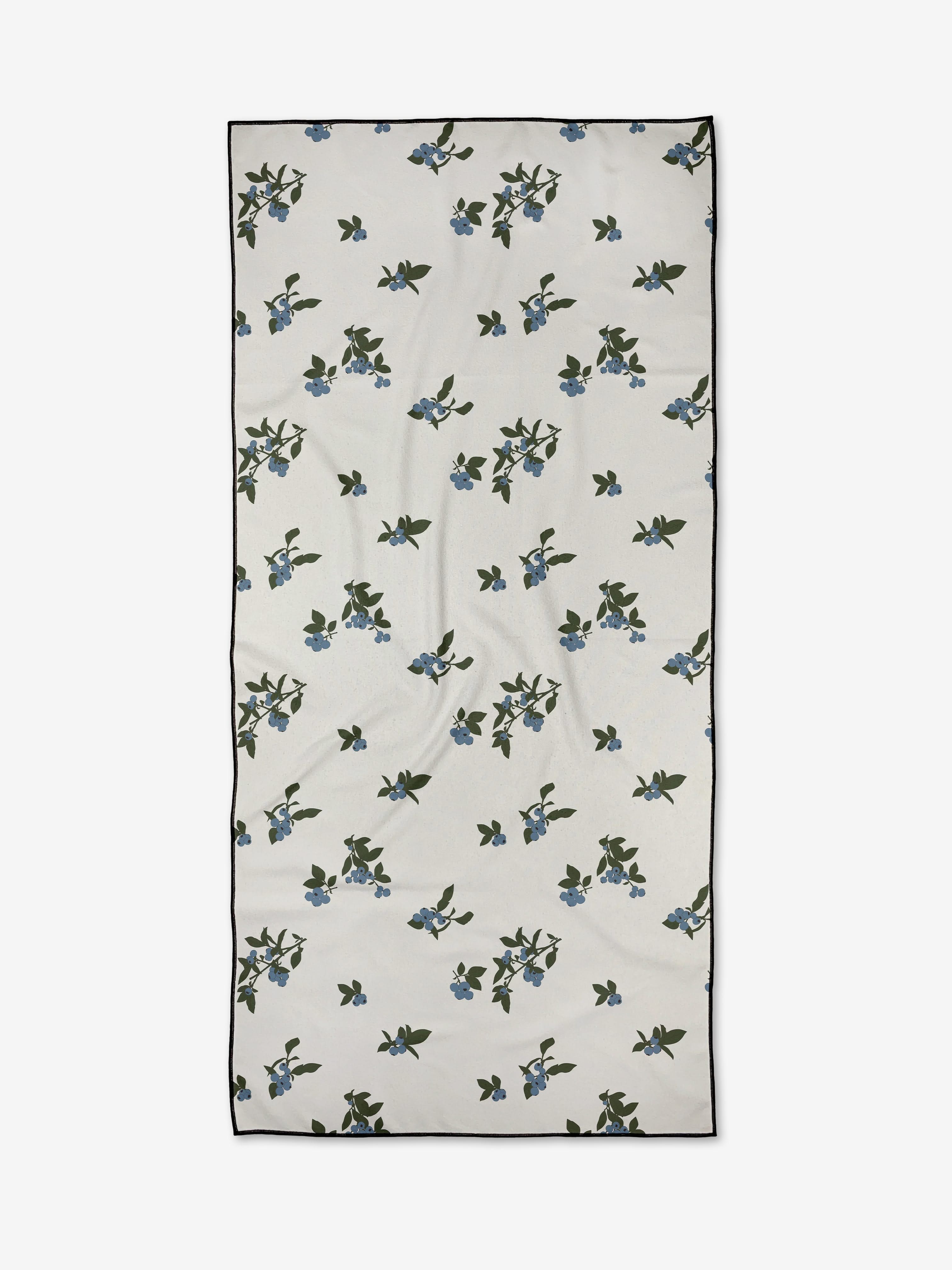 Aubrey Fairchild’s Blueberries is a rectangular white compact beach towel, adorned with scattered green leaves and small blue flowers, finished with a thin black border.