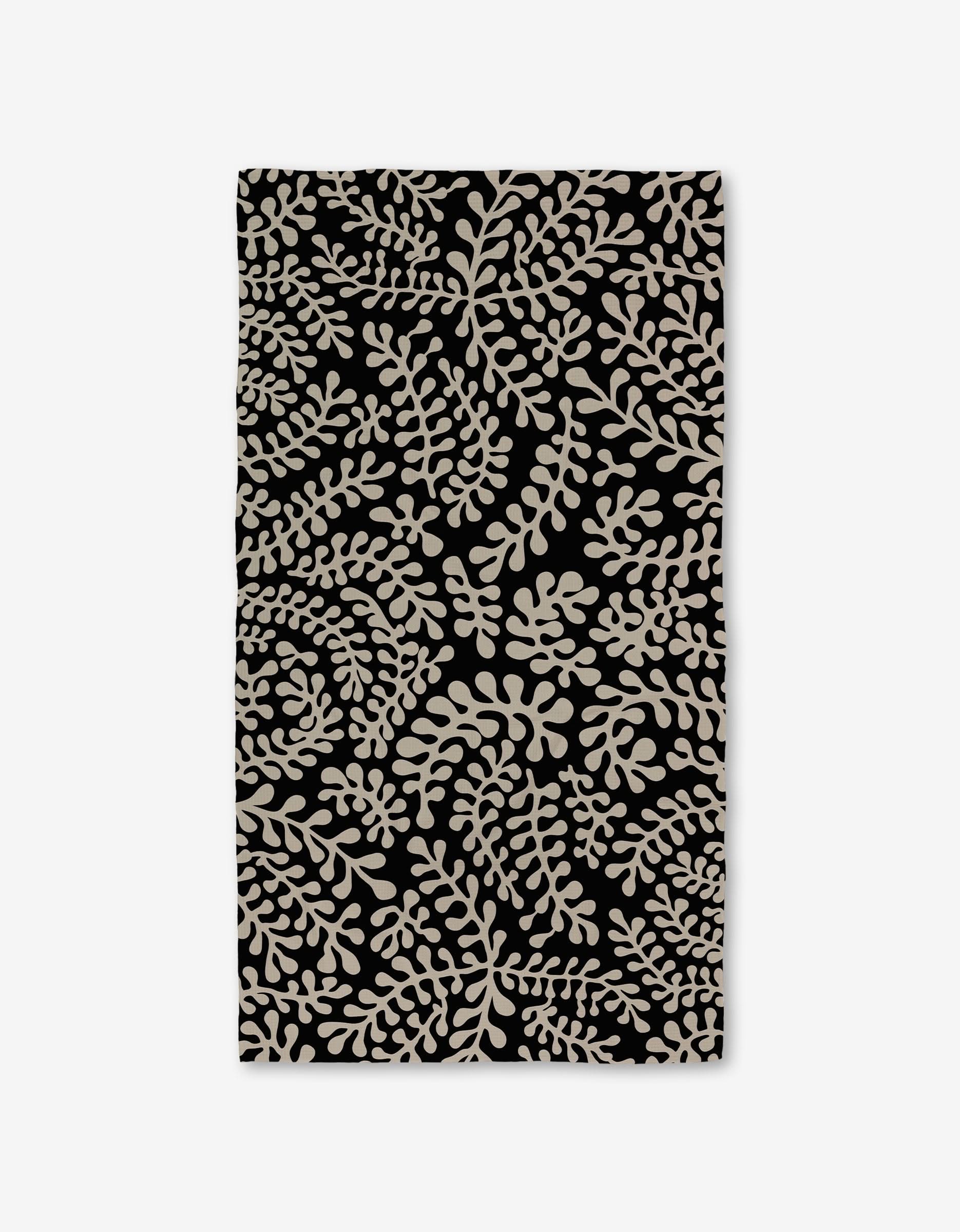 The GEOMETRY Tangled Luxe Bath Towel is rectangular and black, featuring a dense beige leafy pattern. Crafted with Geoweave technology, it offers superior absorbency and softness.