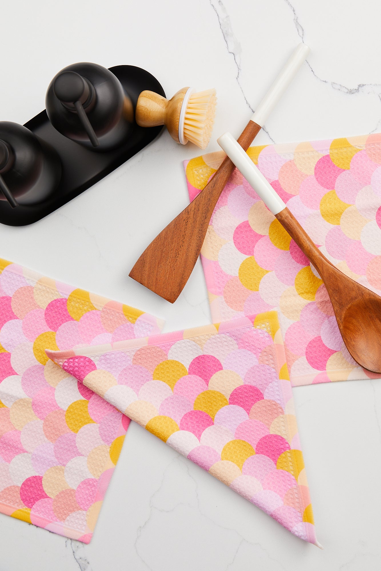 Wooden kitchen utensils, a dish brush, and two black containers sit on a white counter with GEOMETRY Geoweave Inherit dishcloths—one folded and others flat—for a stylish and colorful Dishcloth Set.