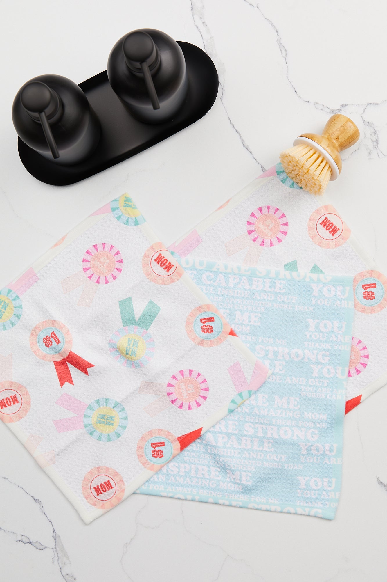 Two GEOMETRY Mom Mantra Geoweave dishcloths, made from recycled materials and featuring colorful designs, sit on a white countertop with a wooden dish brush. Nearby are two black containers for soap or oil on a sleek black tray.