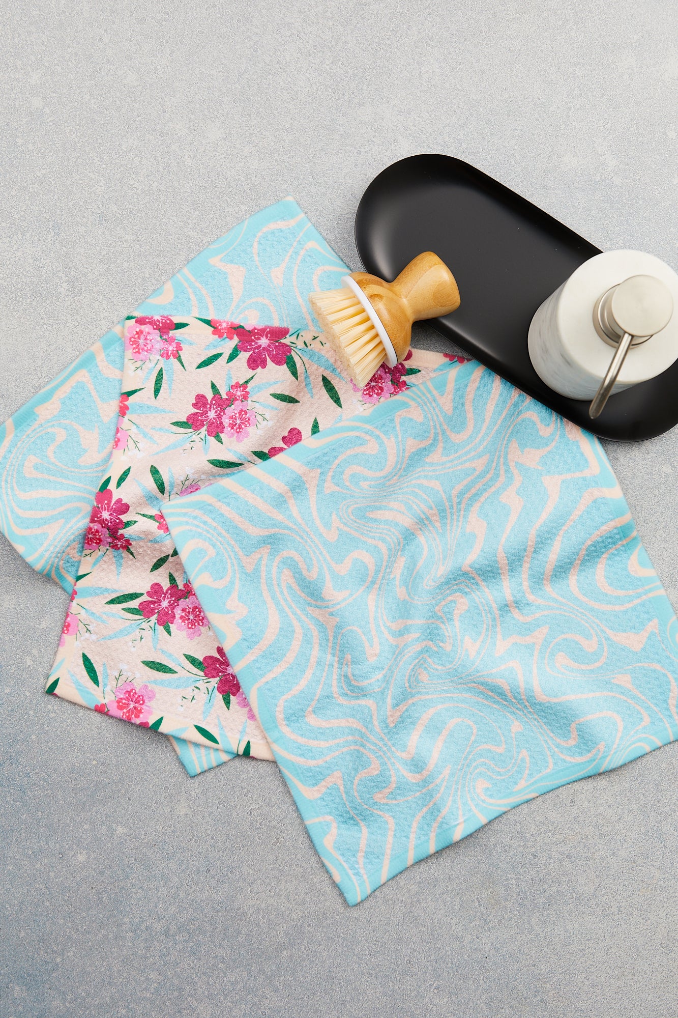 Two GEOMETRY Sending Love Geoweave cleaning cloths—one with pink flowers, one with blue swirls—are shown on a gray surface next to a Dishcloth Set with black tray, wooden brush, and white metal-pump bottle. Super-absorbent and stylish!.