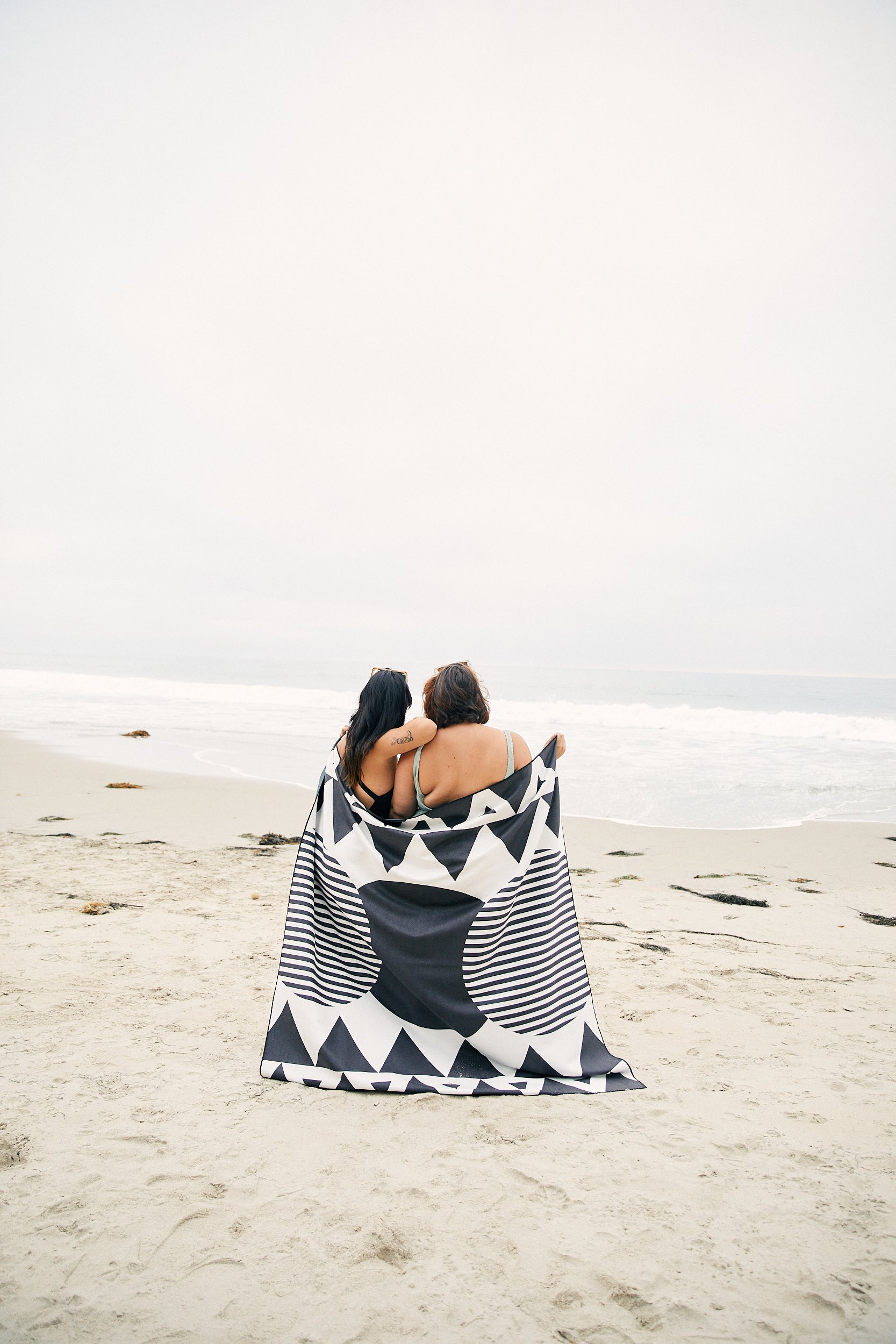 Two people sit side by side on a sandy beach, wrapped together in the GEOMETRY Black Stripes recycled beach blanket. The black and white geometric pattern stands out as they stay cozy by the shore, facing the ocean under an overcast sky.