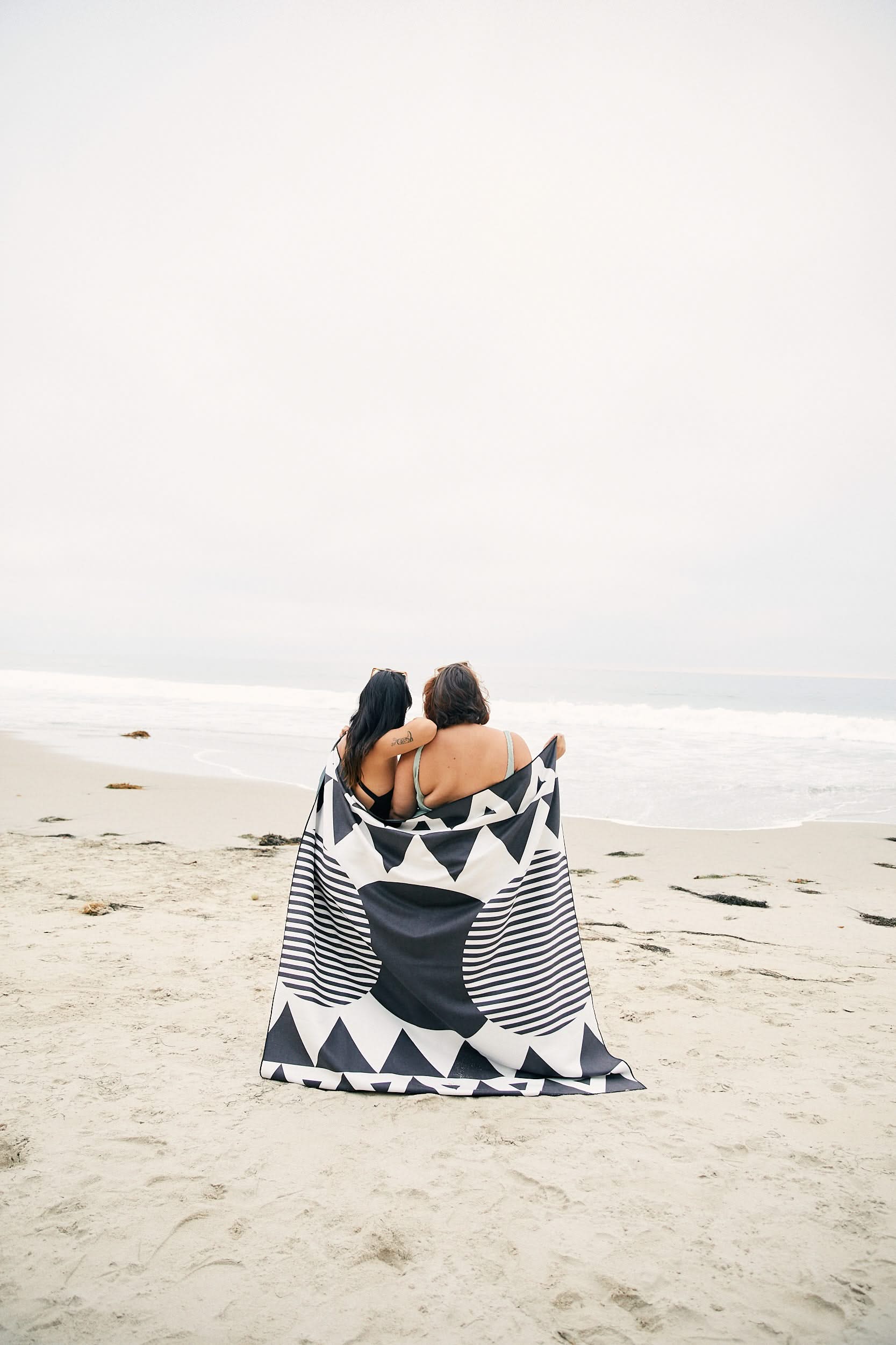 Two people sit closely together on a sandy beach, wrapped in GEOMETRY’s Pink Checkers quick-drying beach blanket, facing the ocean under a cloudy sky.