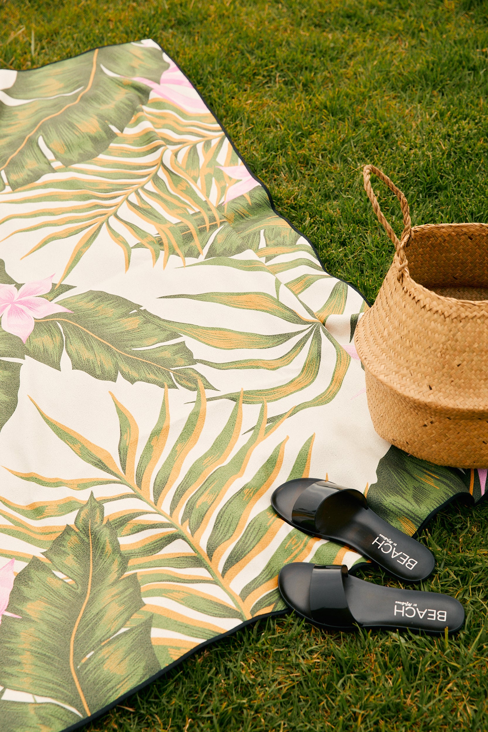 A woven straw bag and black sandals rest beside GEOMETRYs Blue Block Stripes quick-drying beach blanket, made from recycled materials, spread out on green grass.