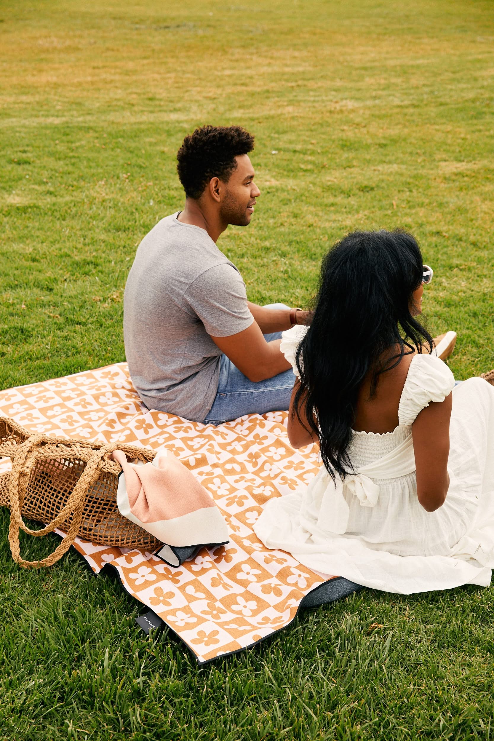 A man in a gray shirt and jeans and a woman in a white dress sit on the GEOMETRY Summer Flowers Beach Blanket on grass. A woven bag and folded blanket are beside them as they face away from the camera.