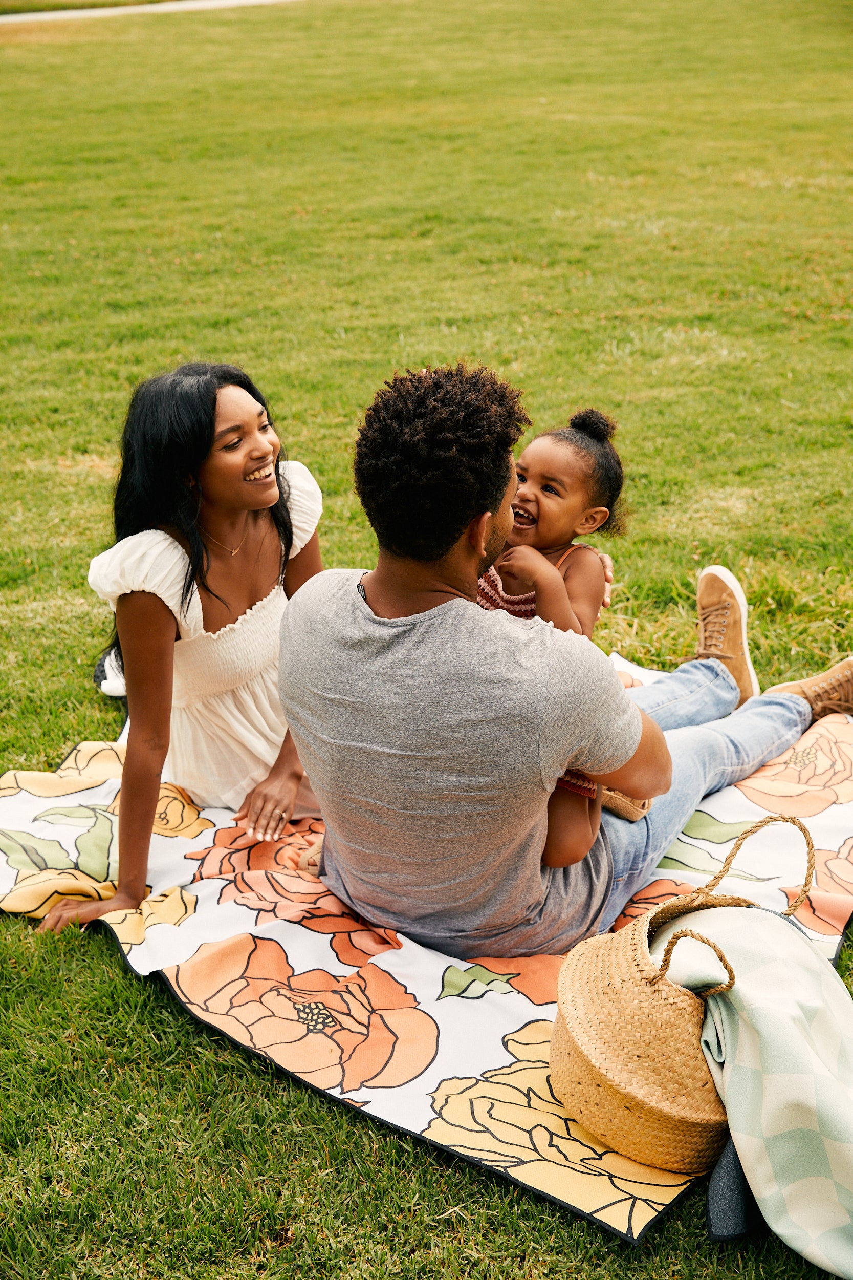 A family enjoys a day at the park on the GEOMETRY Smarties sand-repelling beach blanket. A woman smiles at a man holding a laughing child, with a straw bag nearby.