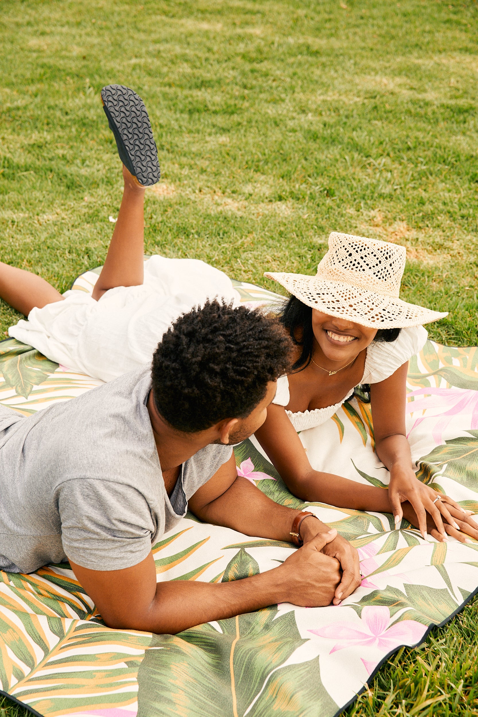 A couple relaxes outdoors on GEOMETRY’s Palm Branches beach blanket, featuring a vibrant tropical pattern and made from recycled materials. The woman, in a sunhat and white dress, smiles beside the man in a gray shirt on the green grass.