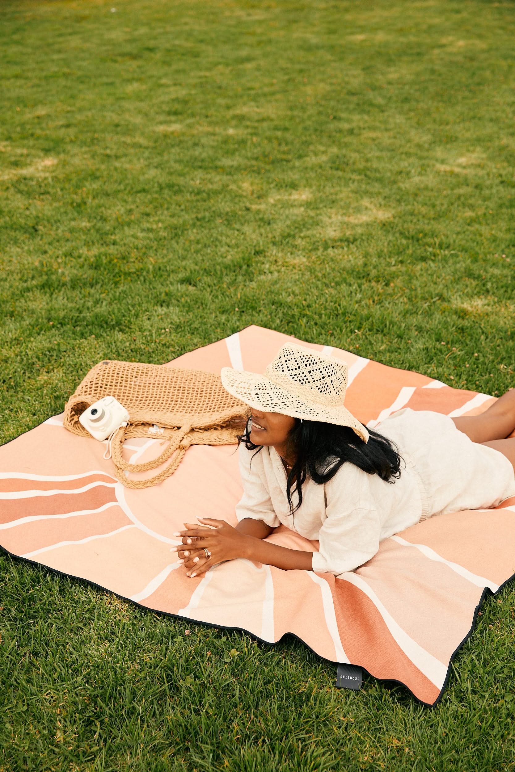 A woman in a sun hat relaxes on GEOMETRY’s Turmeric Pinks recycled beach blanket with a peach and cream pattern, lying on green grass beside her woven bag and camera, enjoying a sunny day outdoors.