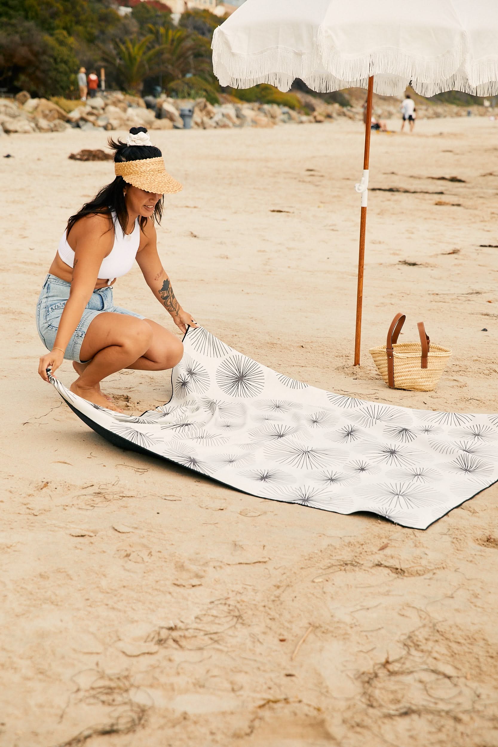 A woman in a straw hat, white crop top, and denim shorts spreads out the GEOMETRY Sea Corals quick-drying Beach Blanket made from recycled materials on sandy beach near a white umbrella and woven basket.