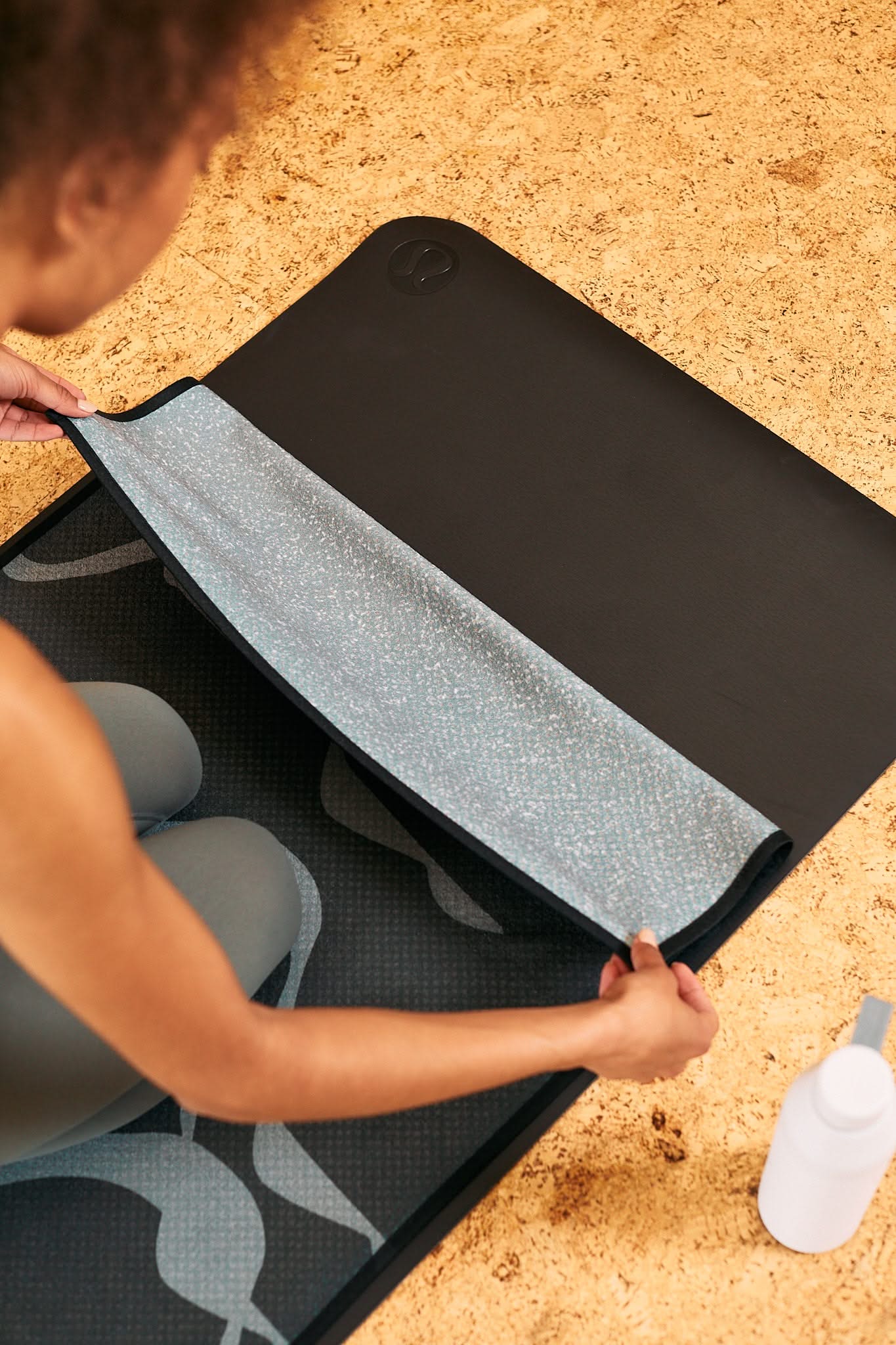 A person kneels on the GEOMETRY Flow yoga mat, lifting one edge to show its silicone grip underside. A water bottle and yoga towel are nearby on the cork floor.