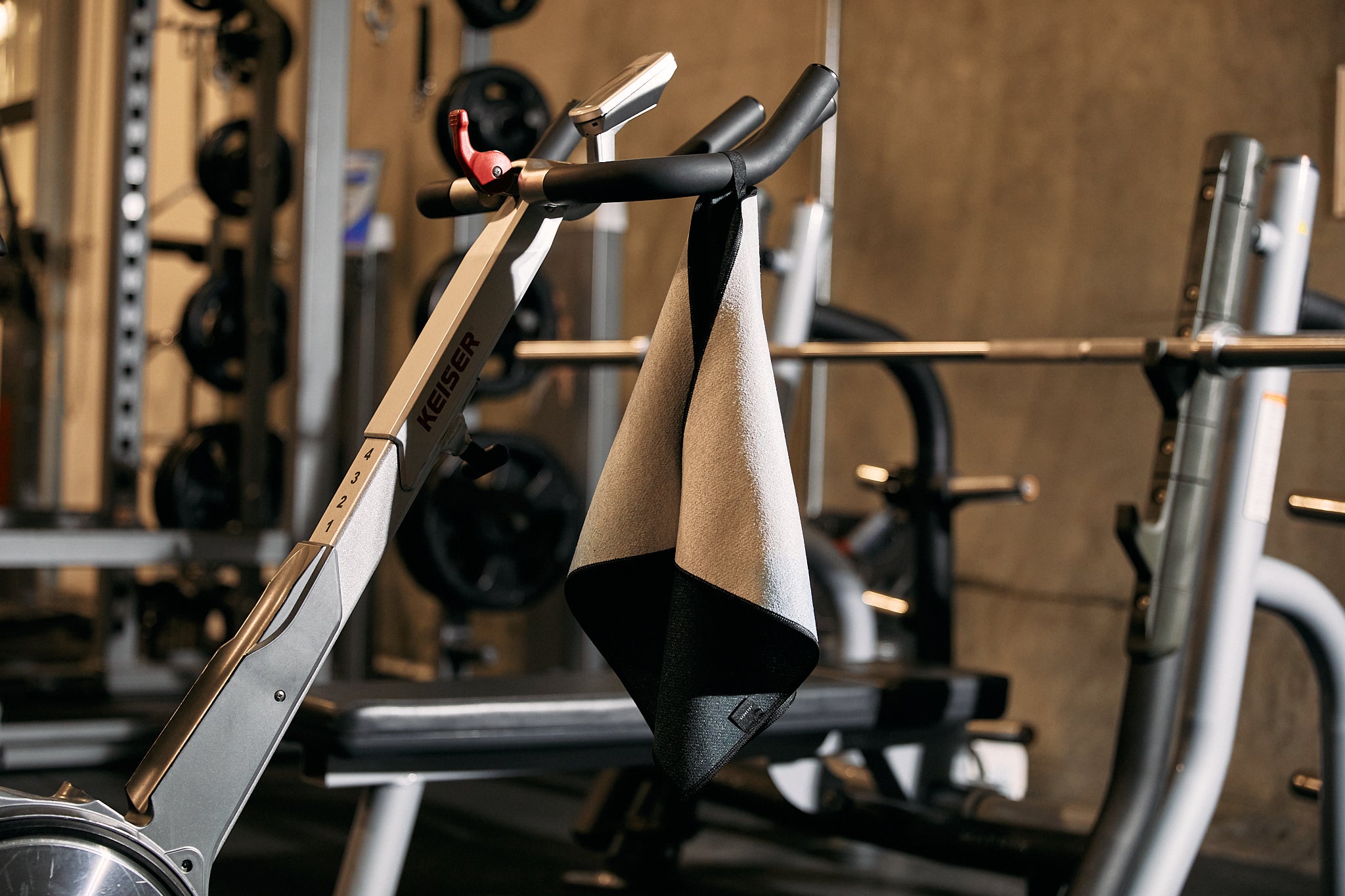 A close-up of a GEOMETRY Root stationary exercise bike with an ultra-absorbent Fitness Towel draped over its handlebars in a gym, with weightlifting equipment visible in the background.