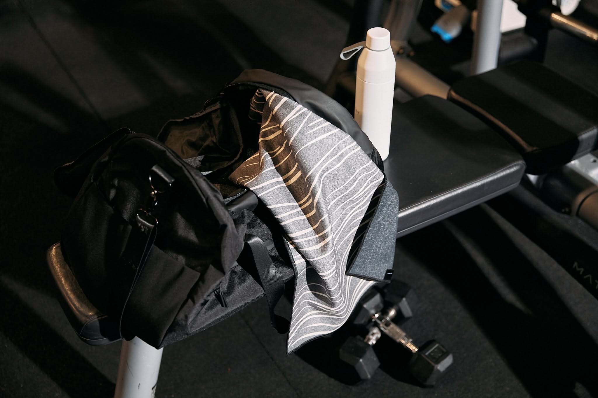 A GEOMETRY Pose ultra-absorbent, fast-drying patterned towel sits on a gym bench with a black bag and white water bottle, while two dumbbells rest nearby under gym lights.