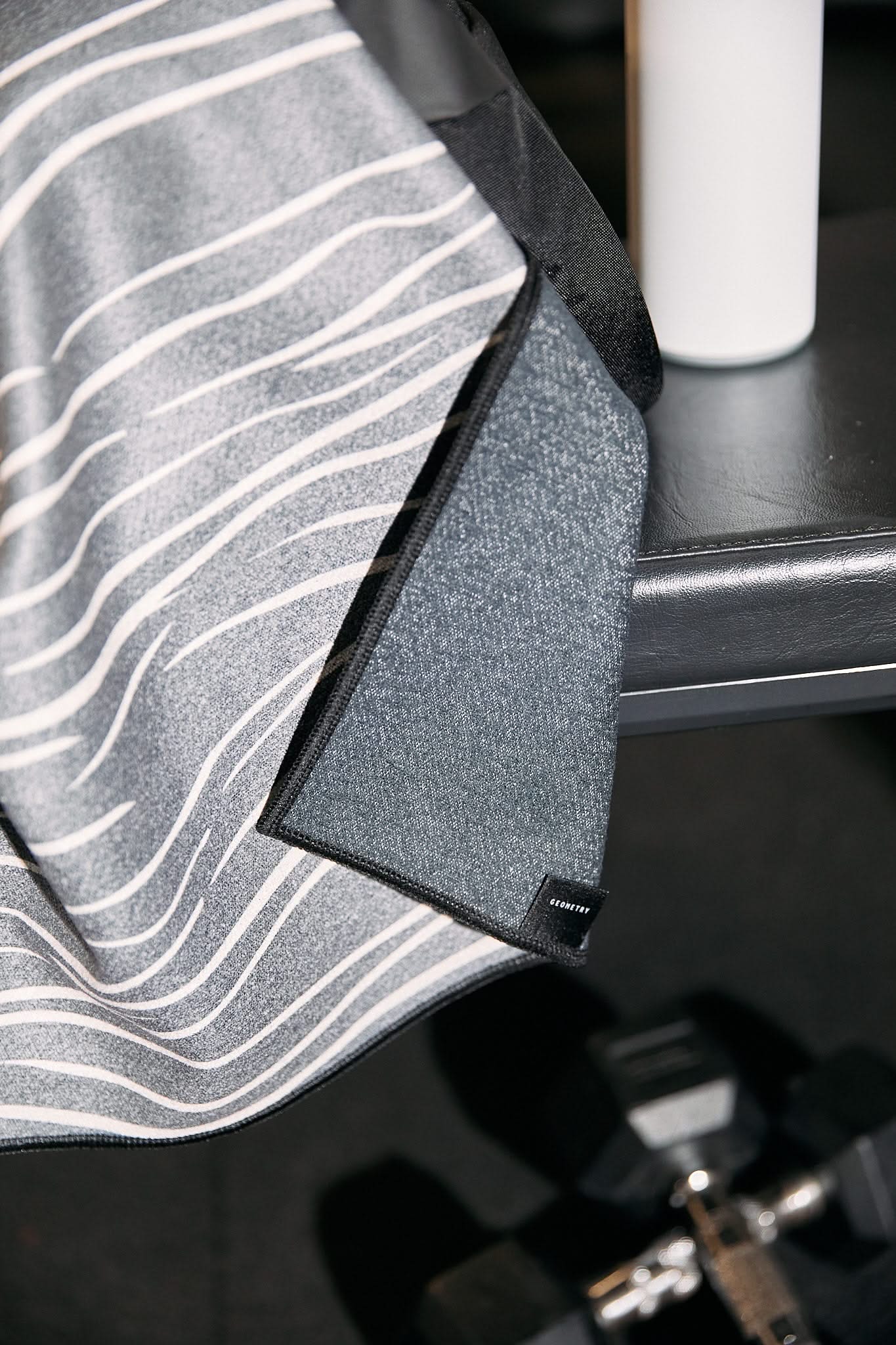 The GEOMETRY Top Layer ultra-absorbent gray and white striped fitness towel is draped over a gym bench, accompanied by a white water bottle on the bench and black dumbbells placed on the floor nearby.
