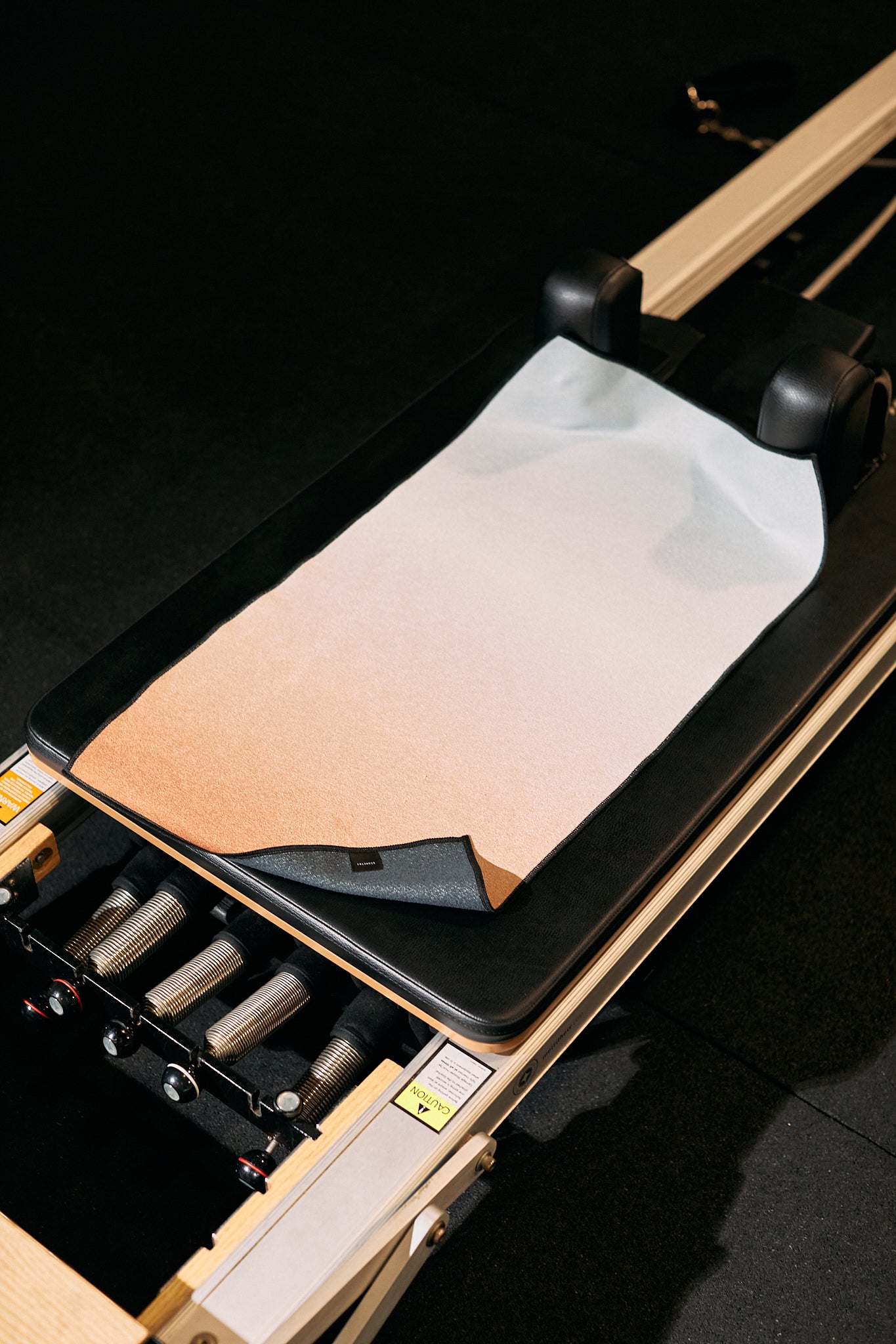 A close-up of GEOMETRYs The Soul, an ultra-absorbent fitness towel made from recycled materials, draped over a Pilates reformer carriage with focus on the springs and black padding.