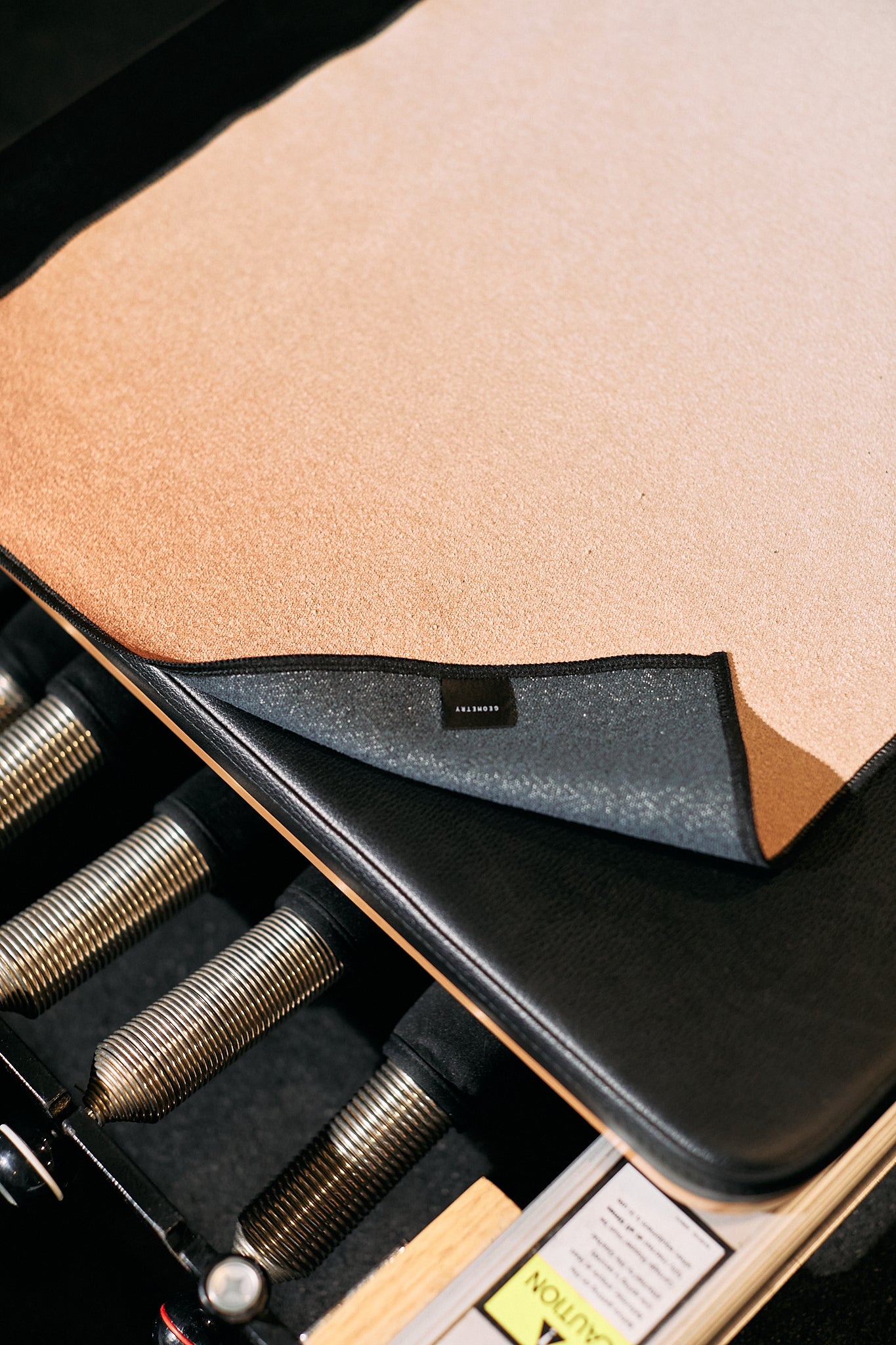 A close-up of the GEOMETRY Where tan exercise mat with a black edge, made from recycled materials, partially folded to show its label and textured underside. The mat lies on fitness equipment featuring metal springs and a caution label.