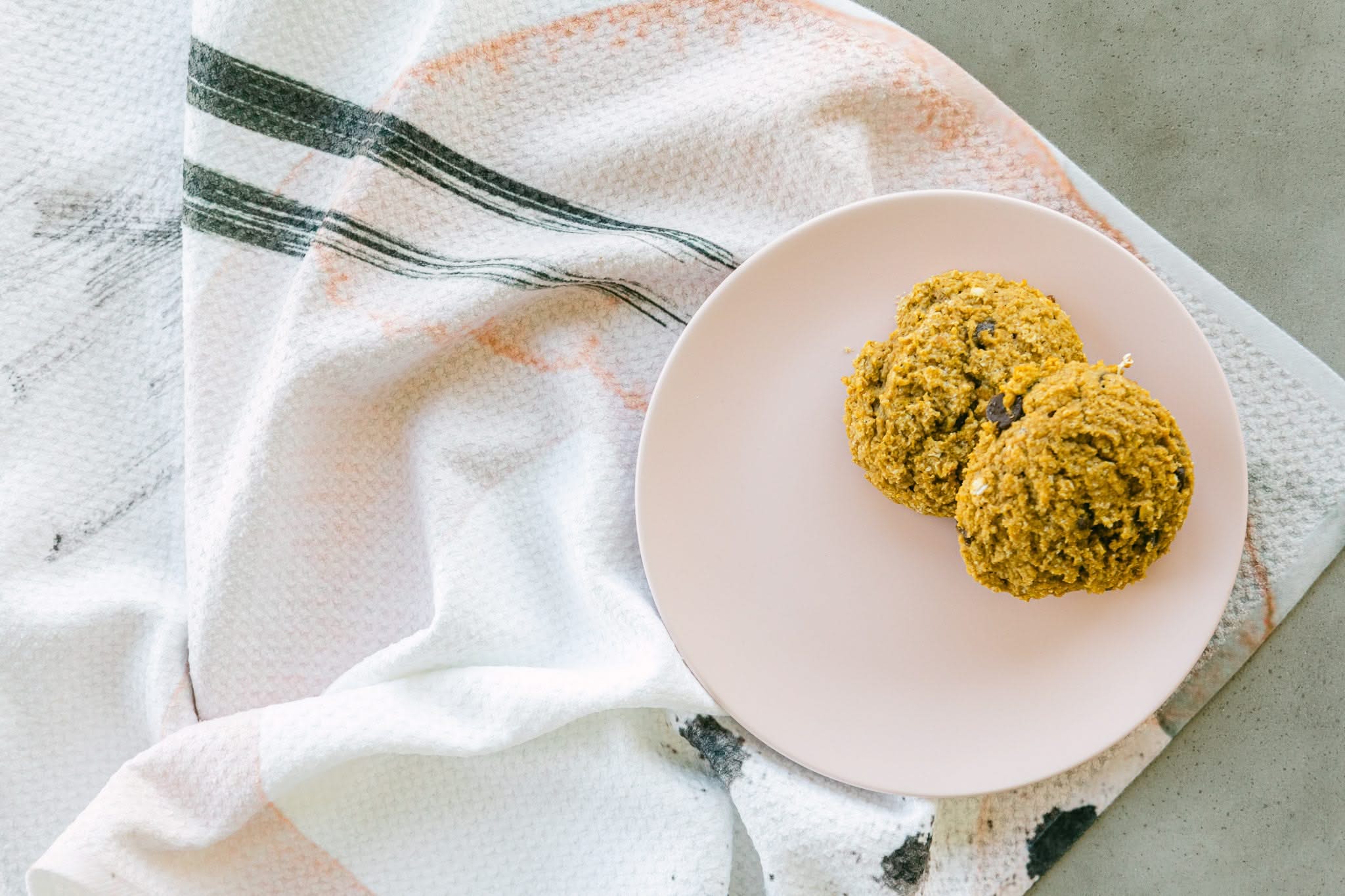 Two oatmeal chocolate chip cookies on a pale pink plate rest atop the Kailee Wright Harper Abstract Tea Towel, featuring black and pink lines, set on a light gray surface.