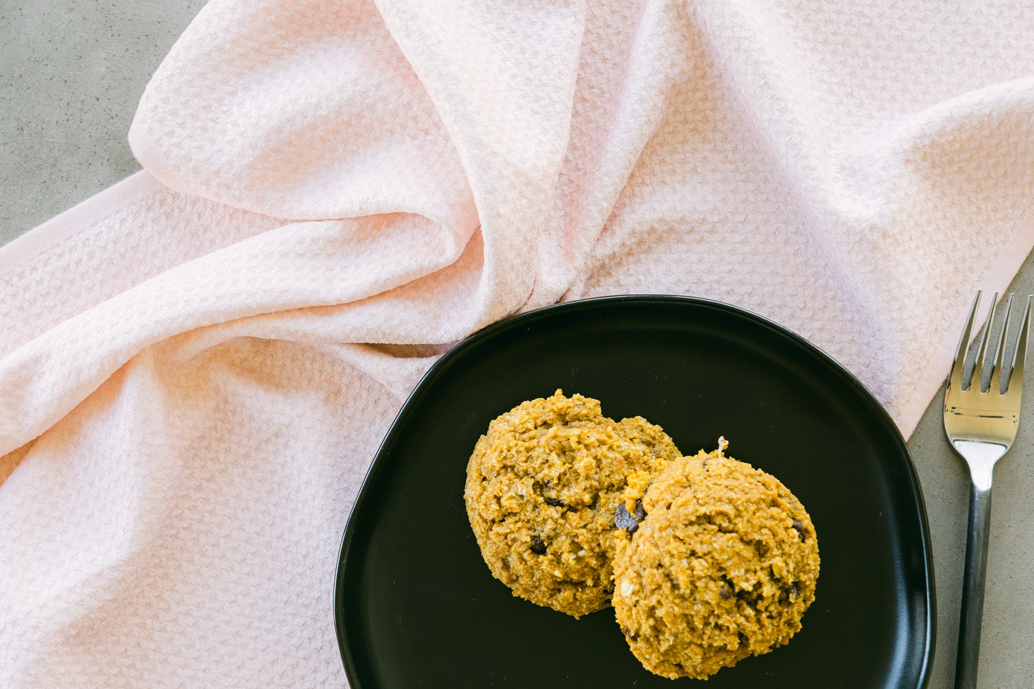 Two chocolate chip cookies sit on a black plate with a silver fork beside the Harper Pink Geoweave dishcloth, all arranged on a gray surface.