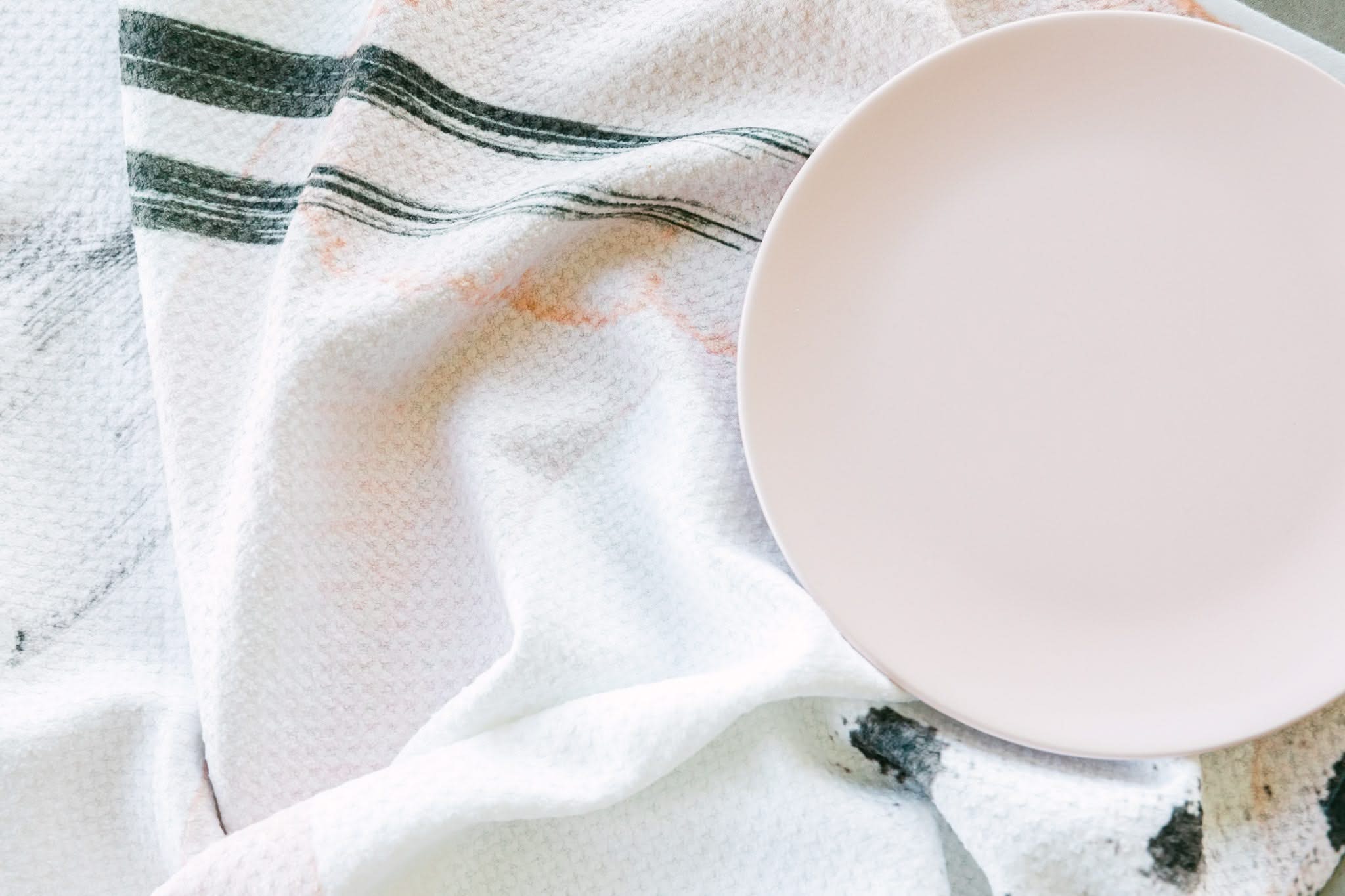 A pale pink Harper Abstract plate by Kailee Wright sits mostly empty on a textured white bar towel with subtle black and peach stripes, resting in the upper right corner. Both items are crafted from recycled materials.