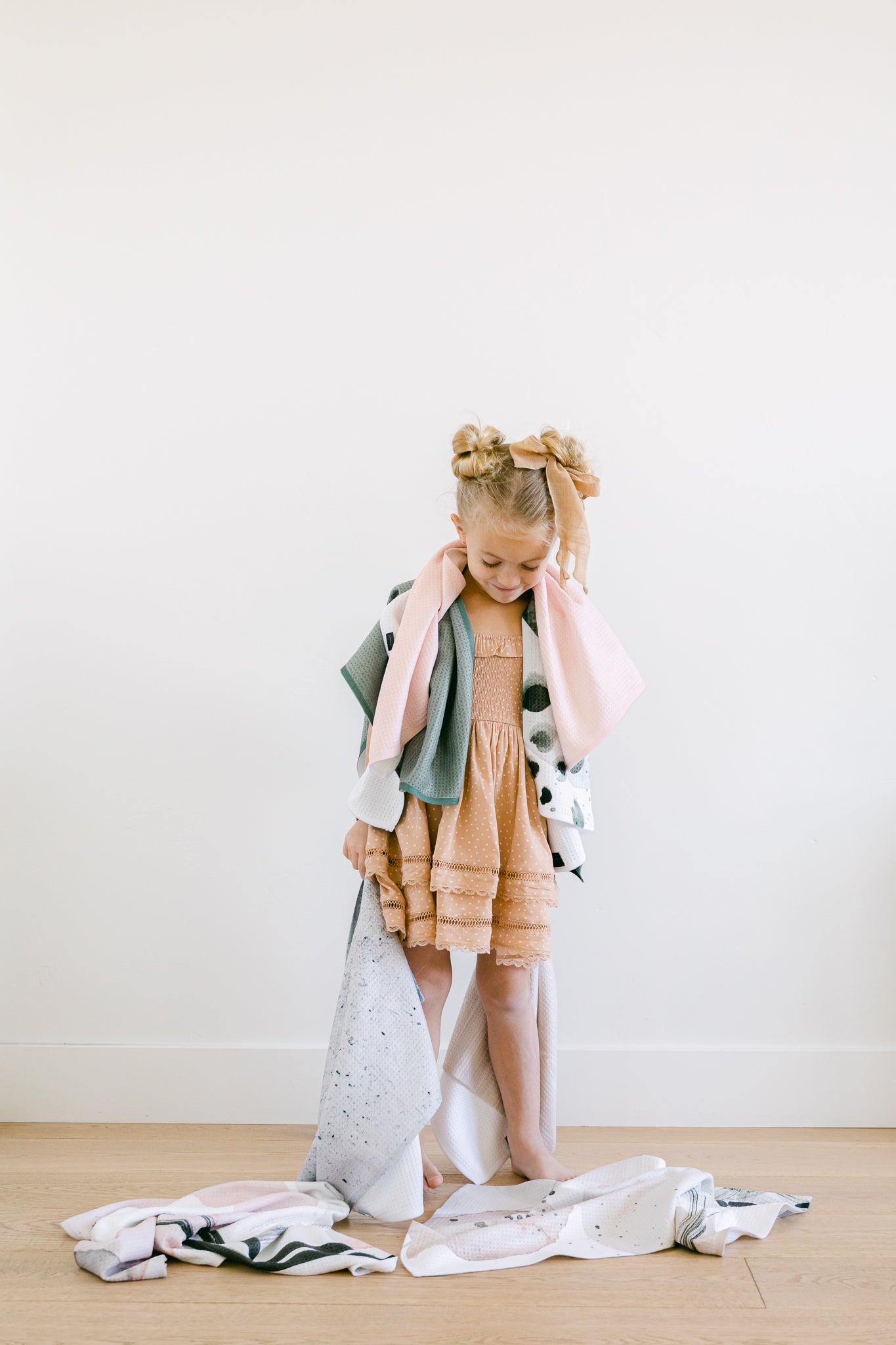 A young girl in a peach dress stands barefoot on a wooden floor, draped with several Kailee Wright Harper Pink Geoweave™ blankets—some wrapped around her shoulders, others trailing on the floor.