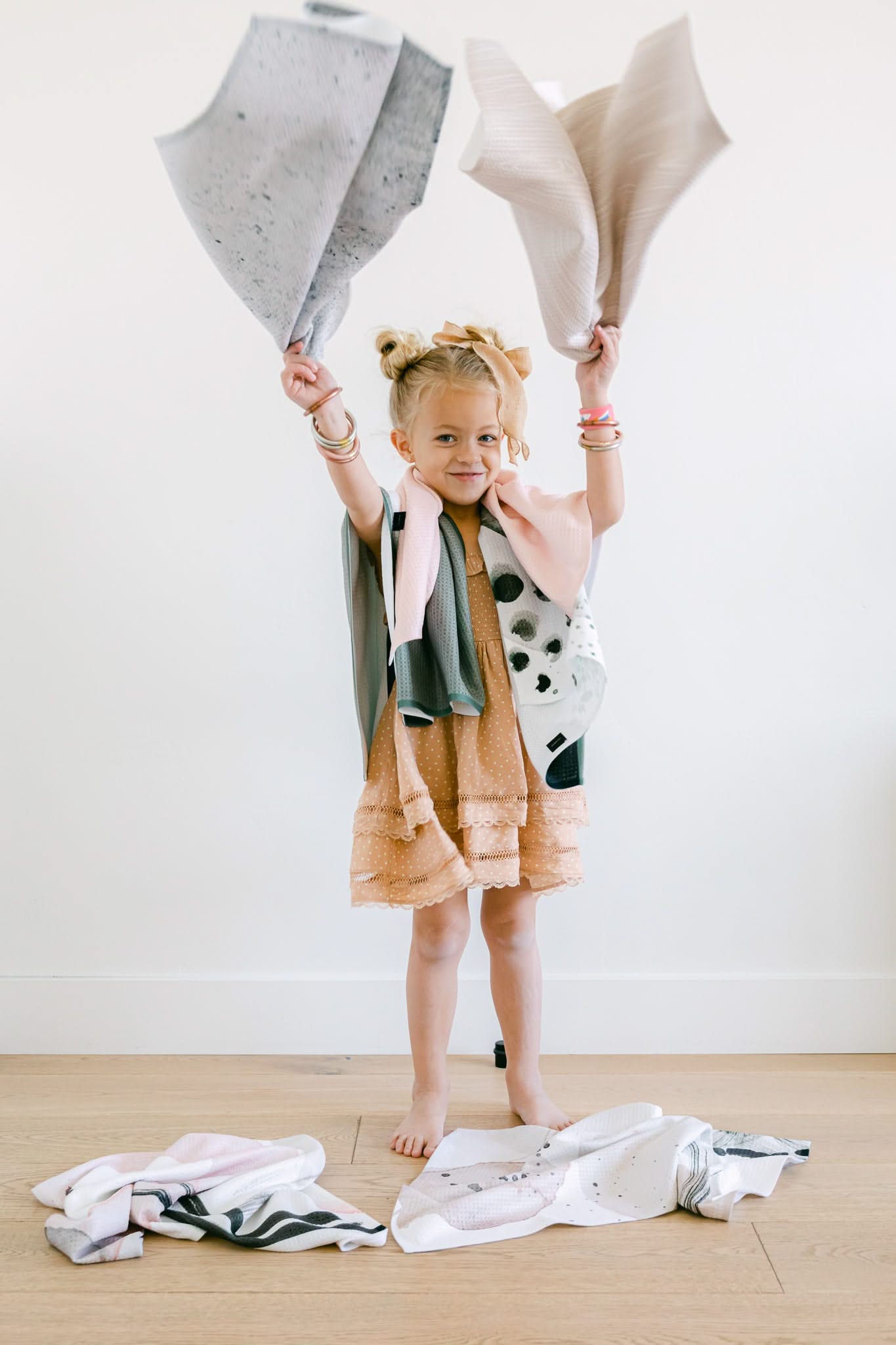 A young girl in a peach dress stands barefoot on a wooden floor, smiling and holding two Kailee Wright Hayden Abstract cloths above her head, with more Hayden Abstract towels at her feet against a white wall.