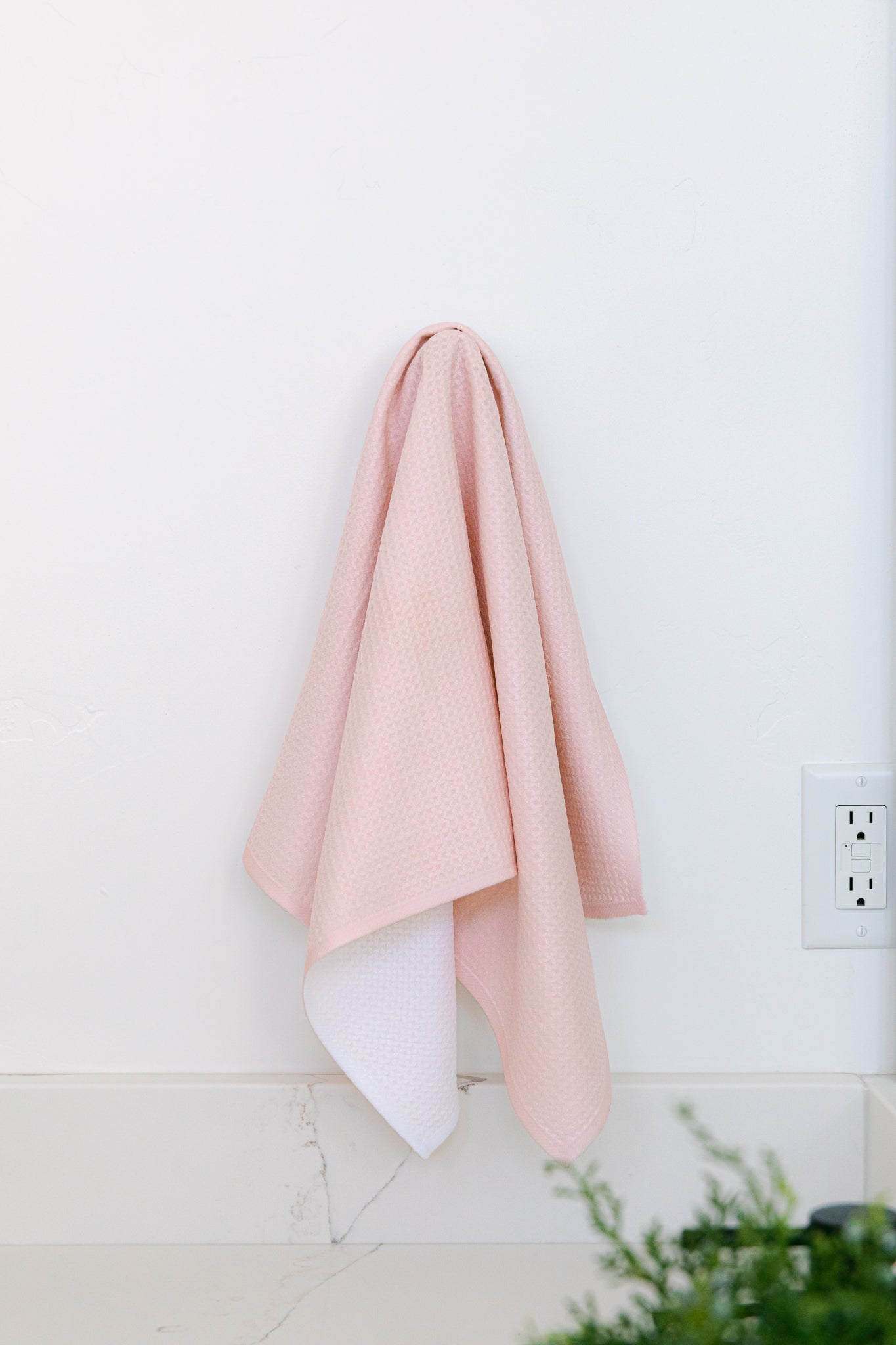 The Harper Pink tea towel by Kailee Wright, crafted from ultra-absorbent Geoweave™ fabric, hangs on a white wall above a baseboard near an outlet, with green foliage partially visible in the bottom right corner.