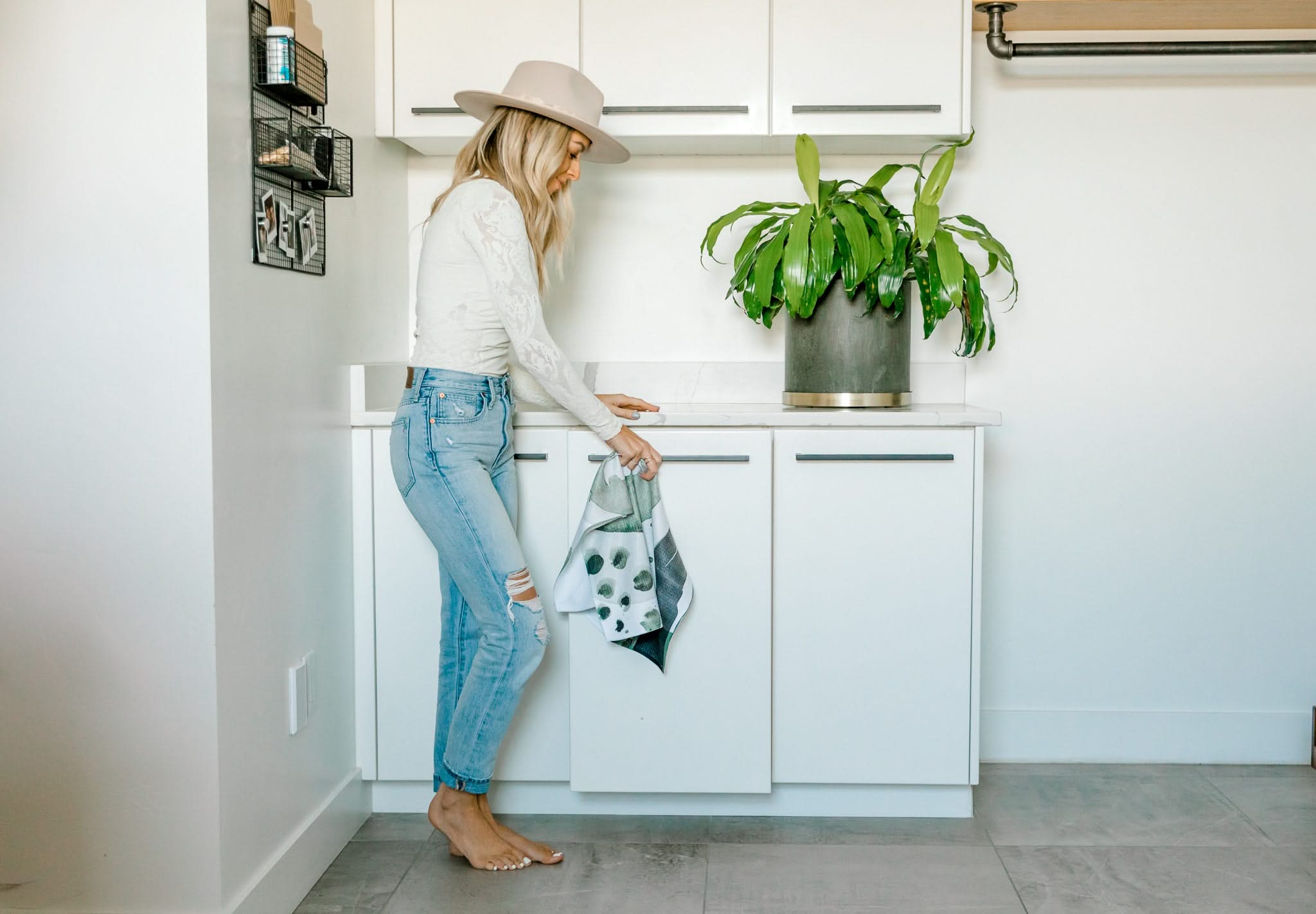 A woman in a hat, white top, and ripped jeans stands barefoot in a modern kitchen, holding a black-and-white reusable bag near a countertop with a potted plant and the Kailee Wright Hunter Abstract tea towel.