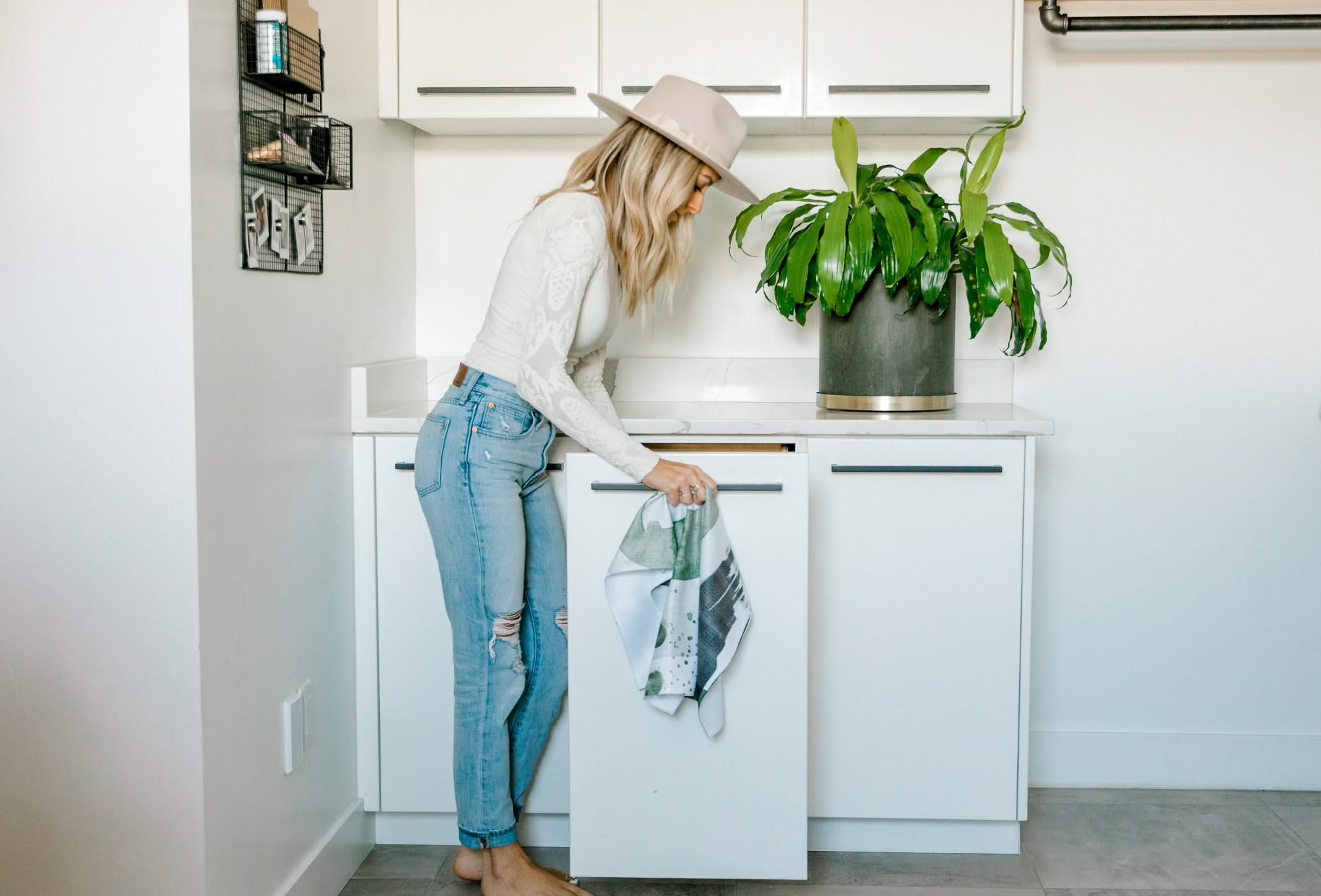 A woman in a hat and jeans opens a kitchen cabinet while holding the Hunter Abstract bar towel by Kailee Wright, with a large potted plant on the white countertop beside her.