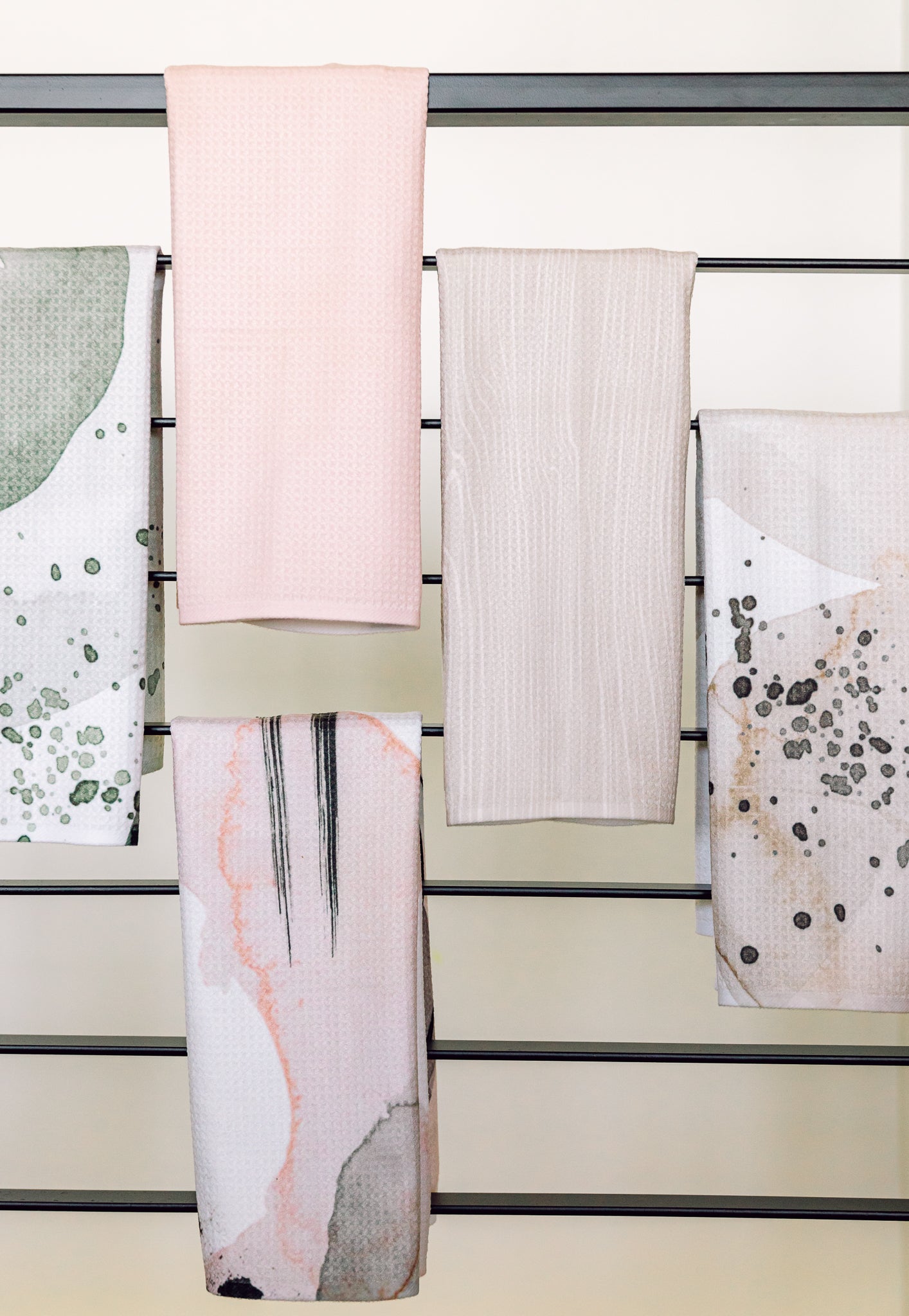 Five Houston Wood towels by Kailee Wright hang on a horizontal rack—two in solid pastel pink and beige, three with abstract green, pink, and gray patterns. Made from recycled materials, they add eco-friendly style to the light wall behind.