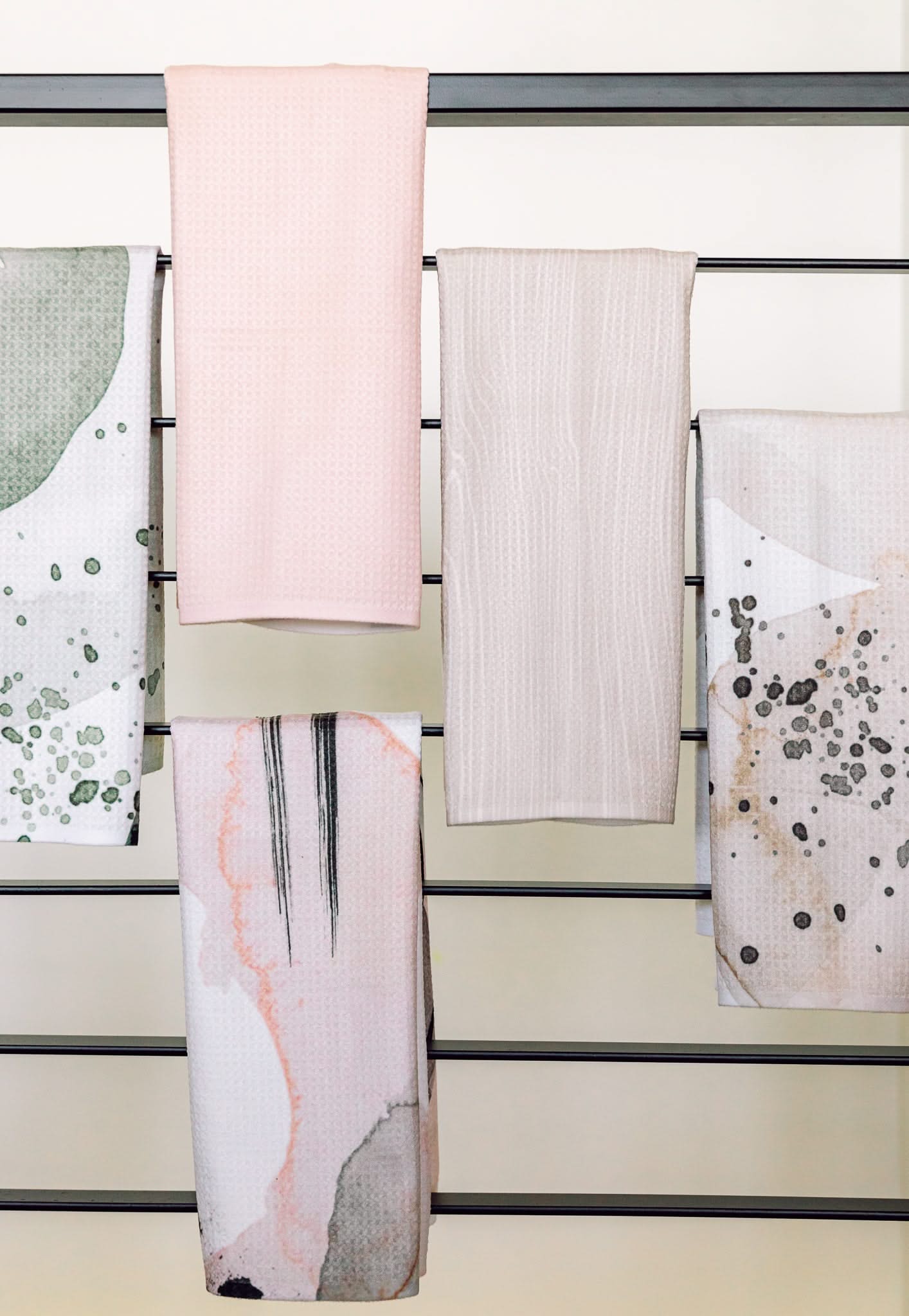 Five Houston Wood towels by Kailee Wright hang on a horizontal rack—two in solid pastel pink and beige, three with abstract green, pink, and gray patterns. Made from recycled materials, they add eco-friendly style to the light wall behind.