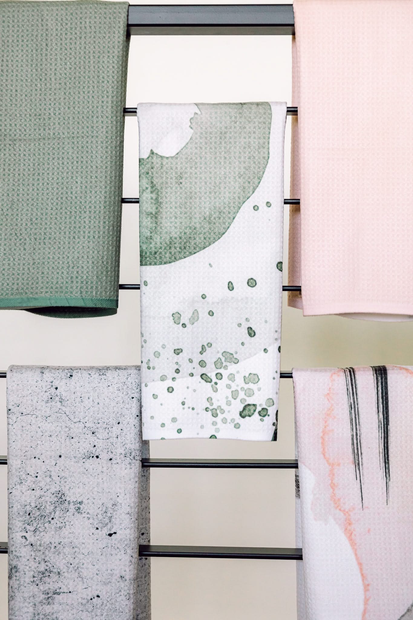 Five Kailee Wright towels, including the ultra-absorbent Hunter Green and a recycled kitchen towel, hang on a rack in pastel patterns—blending style with eco-friendly function.
