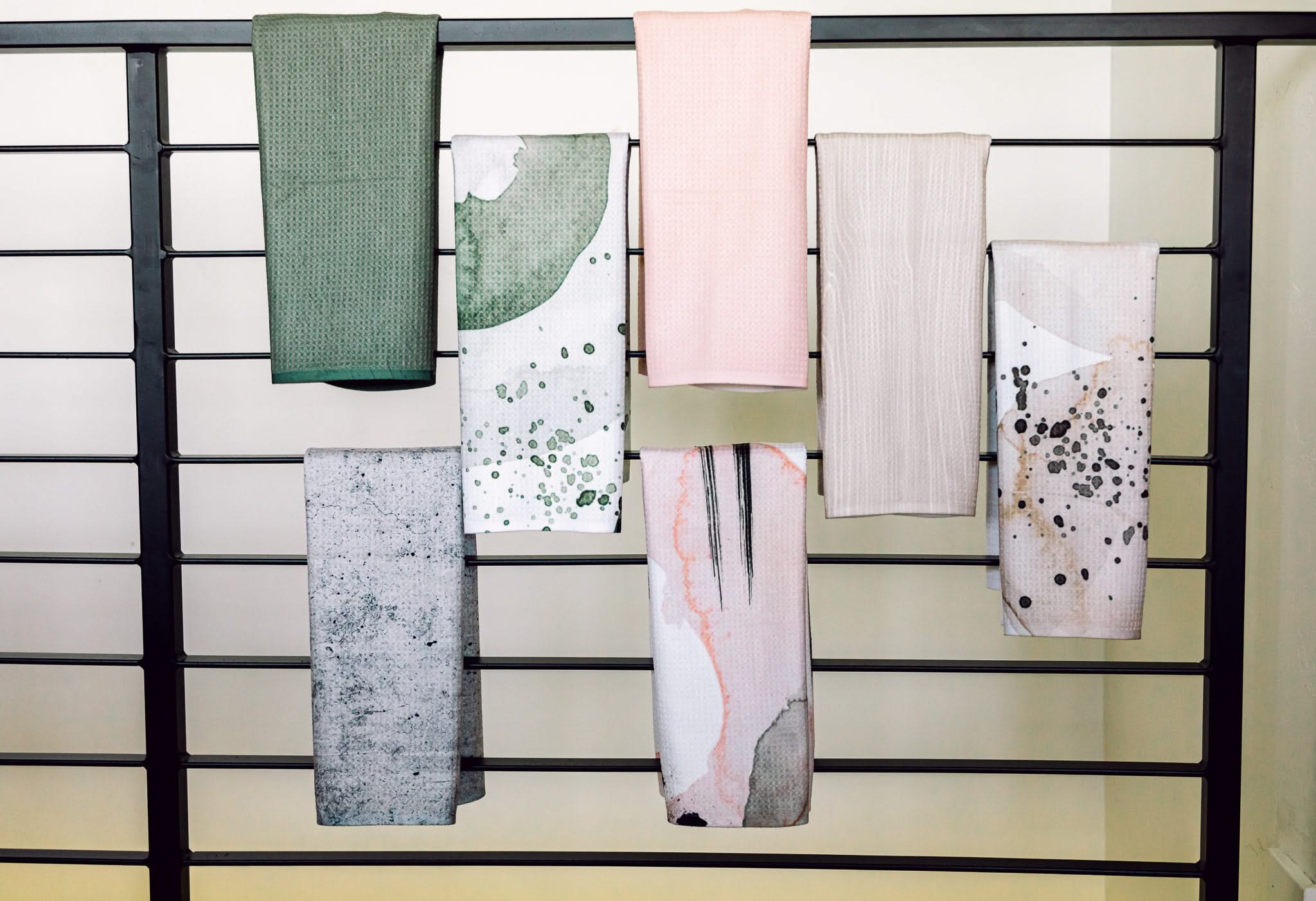 Six patterned and solid-colored towels from Kailee Wrights Hayden Abstract collection, including an ultra-absorbent Geoweave tea towel, hang on a black railing. Designs feature green, pink, beige, gray, and abstract prints against a light wall.