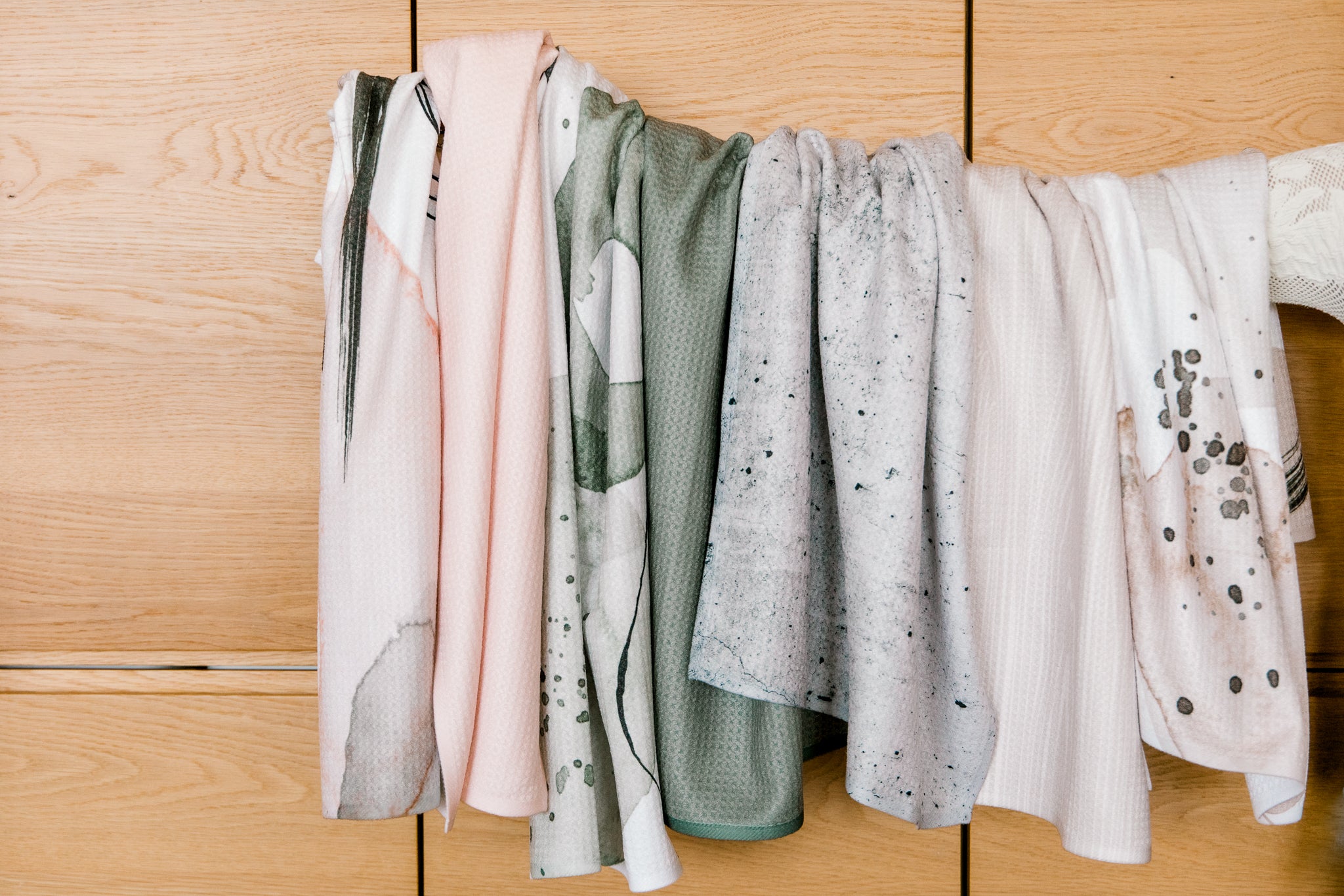 A row of Kailee Wright’s Hudson Grey dishcloths in soft pink, green, white, and grey shades—some crafted from Geoweave fabric or recycled materials—hangs over wood, showcasing abstract and speckled patterns.